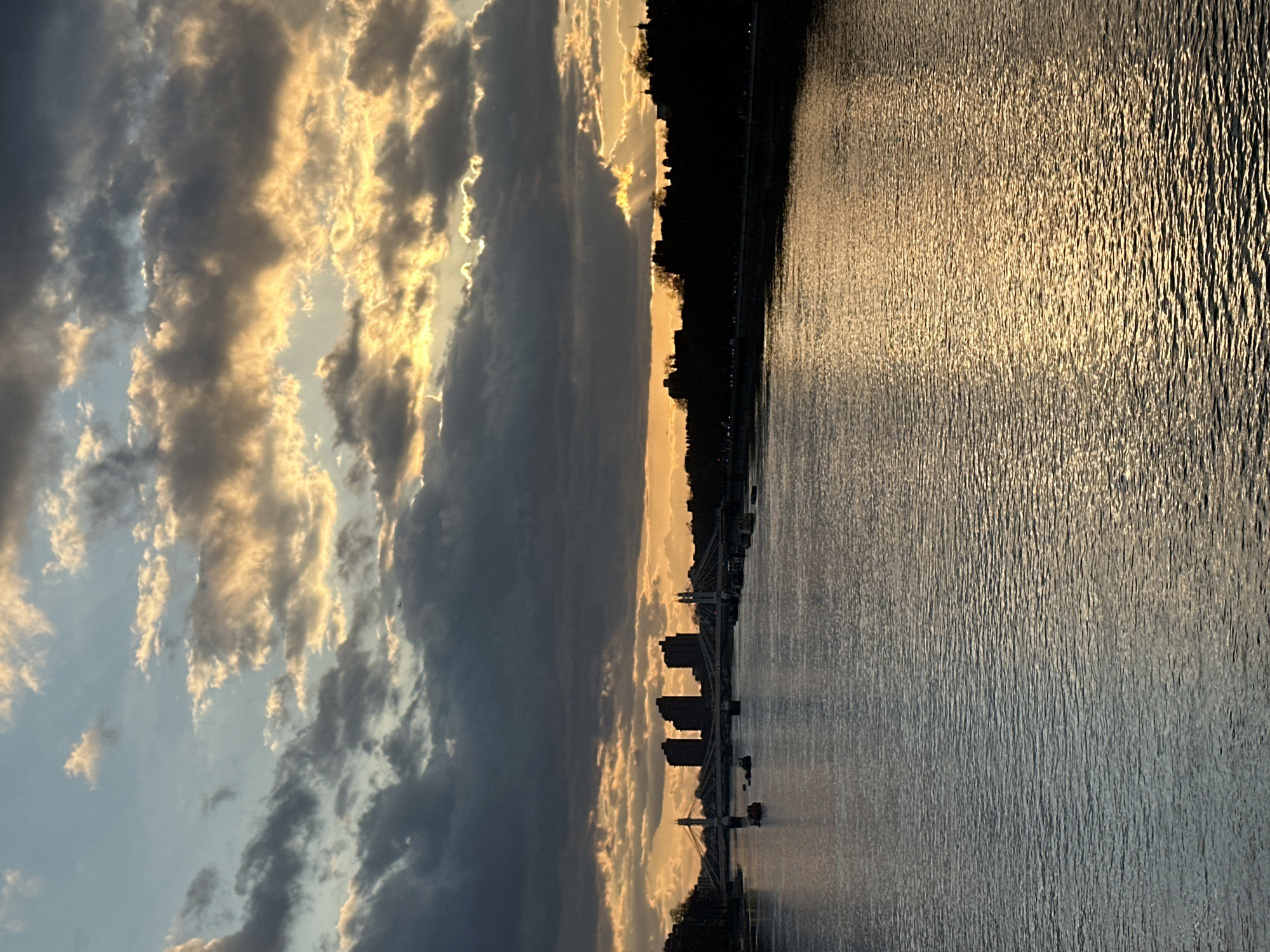 The image shows a waterscape with a cityscape at sunset. The sky is filled with clouds, some dark gray and others illuminated with a golden glow from the setting sun. The river reflects the golden light, creating a shimmering effect on the water surface. A dark tree line runs along the riverbank, with buildings visible in the distance, suggesting a city. Based on the presence of a suspension bridge resembling the Hungerford Bridge and the buildings lining the river, the location can be identified as London, United Kingdom. There are a few boats in the water in front of the cityscape.