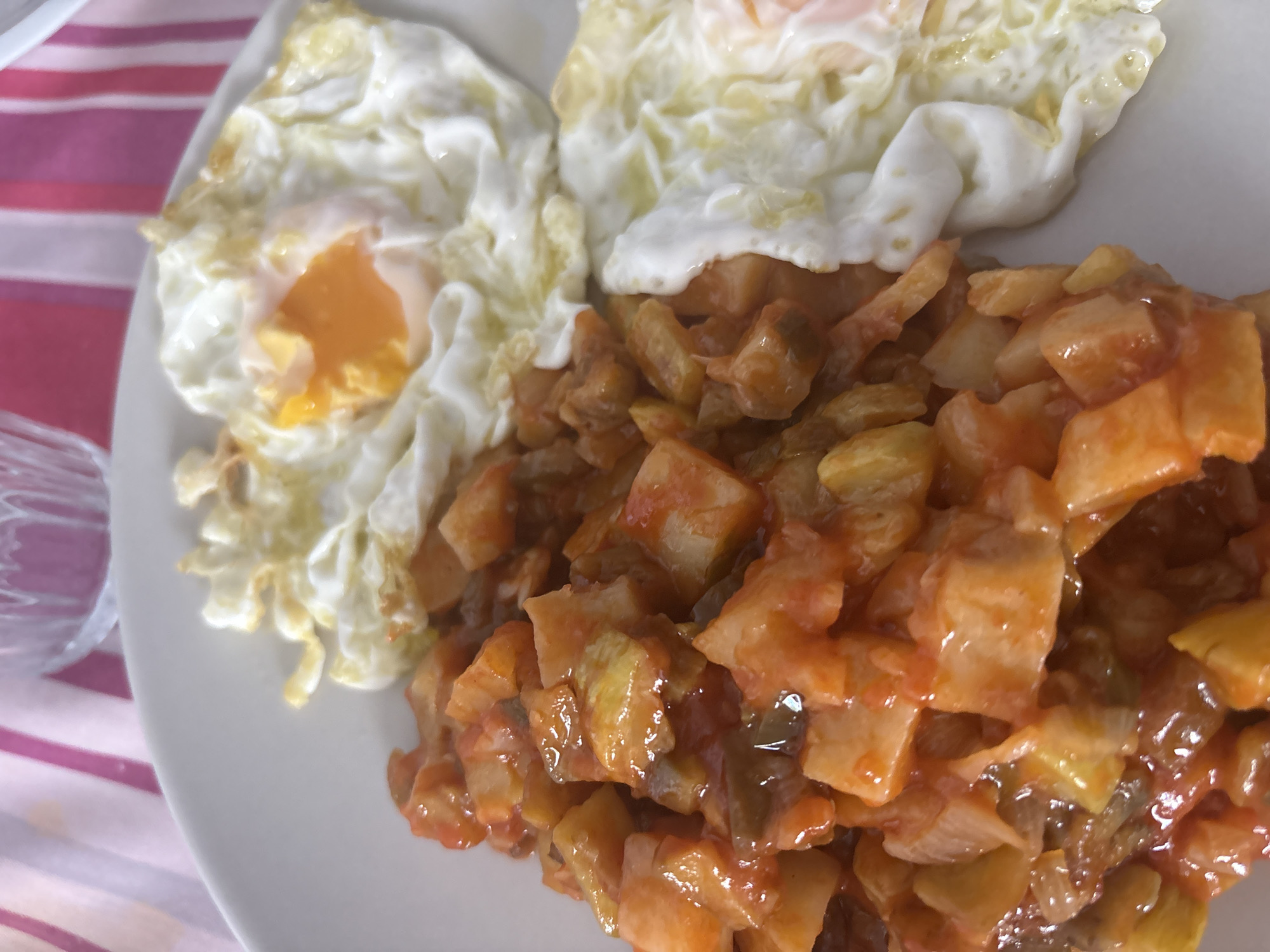 The image presents a directly overhead, slightly angled view of a meal served on a white, round plate. The plate rests on a vertically striped fabric tablecloth, featuring alternating wide magenta and narrow white bands. On the left side of the plate, a portion of diced, cooked vegetables, predominantly yellowish-brown cubes resembling potatoes, is coated in a glossy, reddish-orange sauce, with darker vegetable pieces interspersed. To the right, two fried eggs are visible. The upper egg displays a bright orange, semi-liquid yolk encircled by a crispy, white, and ruffled albumen. The lower egg shows a similar white and ruffled albumen. In the upper left background, a portion of a clear, textured glass is visible out of focus. The setting is an indoor dining environment located in Urbanización Encinares De Alcolea, Spain.