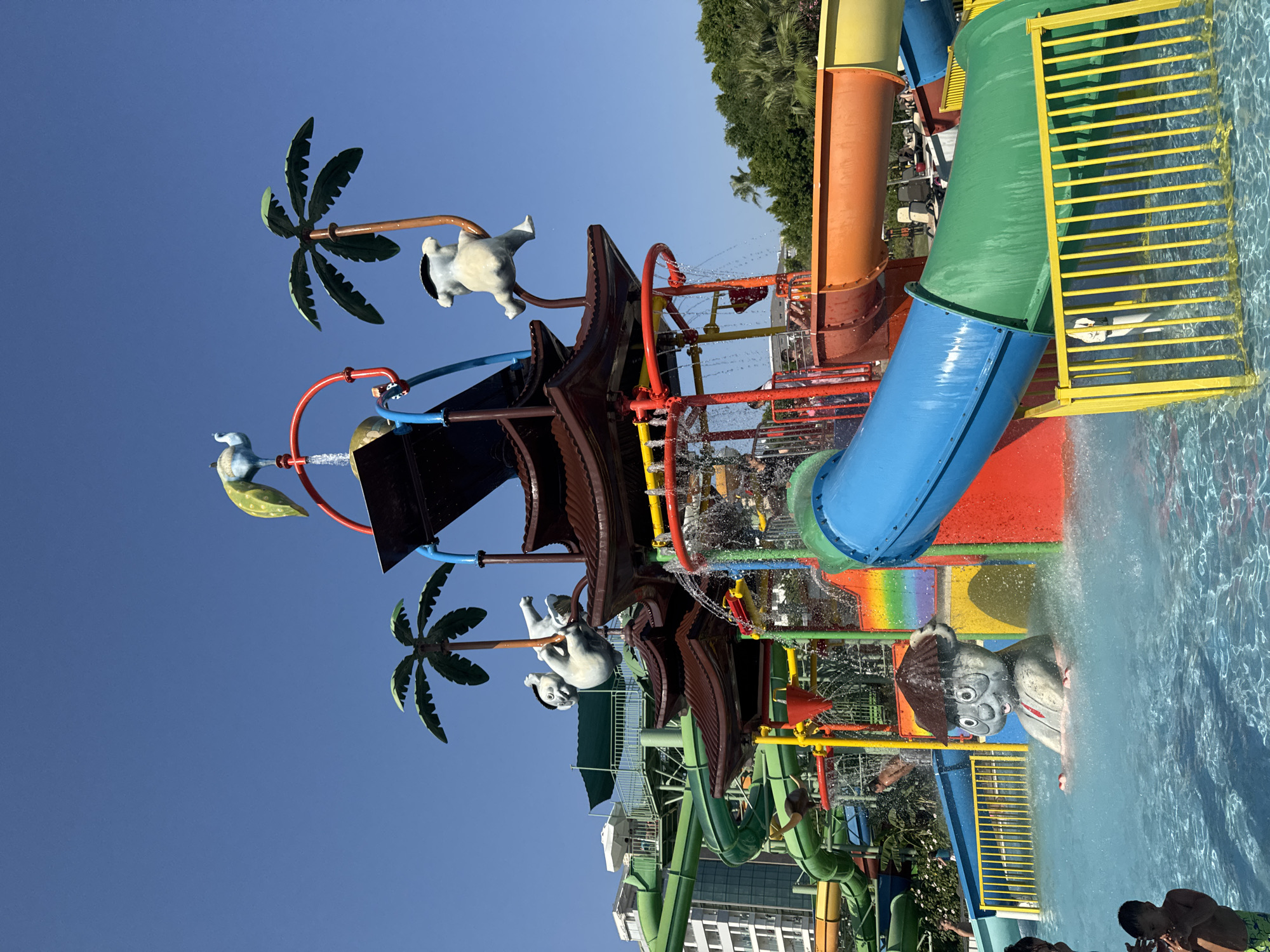 The image depicts a vibrant water park under a clear blue sky. The central feature is a multi-level water slide structure with a pagoda-like roof. Several slides of various colors, including green, blue, orange, and yellow, spiral downwards. Decorative elements like palm trees and cartoonish figures, including a large grey creature with a mushroom-shaped hat sitting in the water, are integrated into the design. Water is actively spraying from various points on the structure, creating a playful atmosphere. In the foreground, two young boys are visible in the shallow pool. In the background, a multi-story building with white balconies and balconies with green awnings suggests the proximity of a resort or hotel. Lush green trees are visible in the far background, and a glimpse of a wider sandy area with chairs and umbrellas hints at more recreational facilities. The location is identified as Aksu, Turkey, which is known for its tourist resorts along the Mediterranean coast.