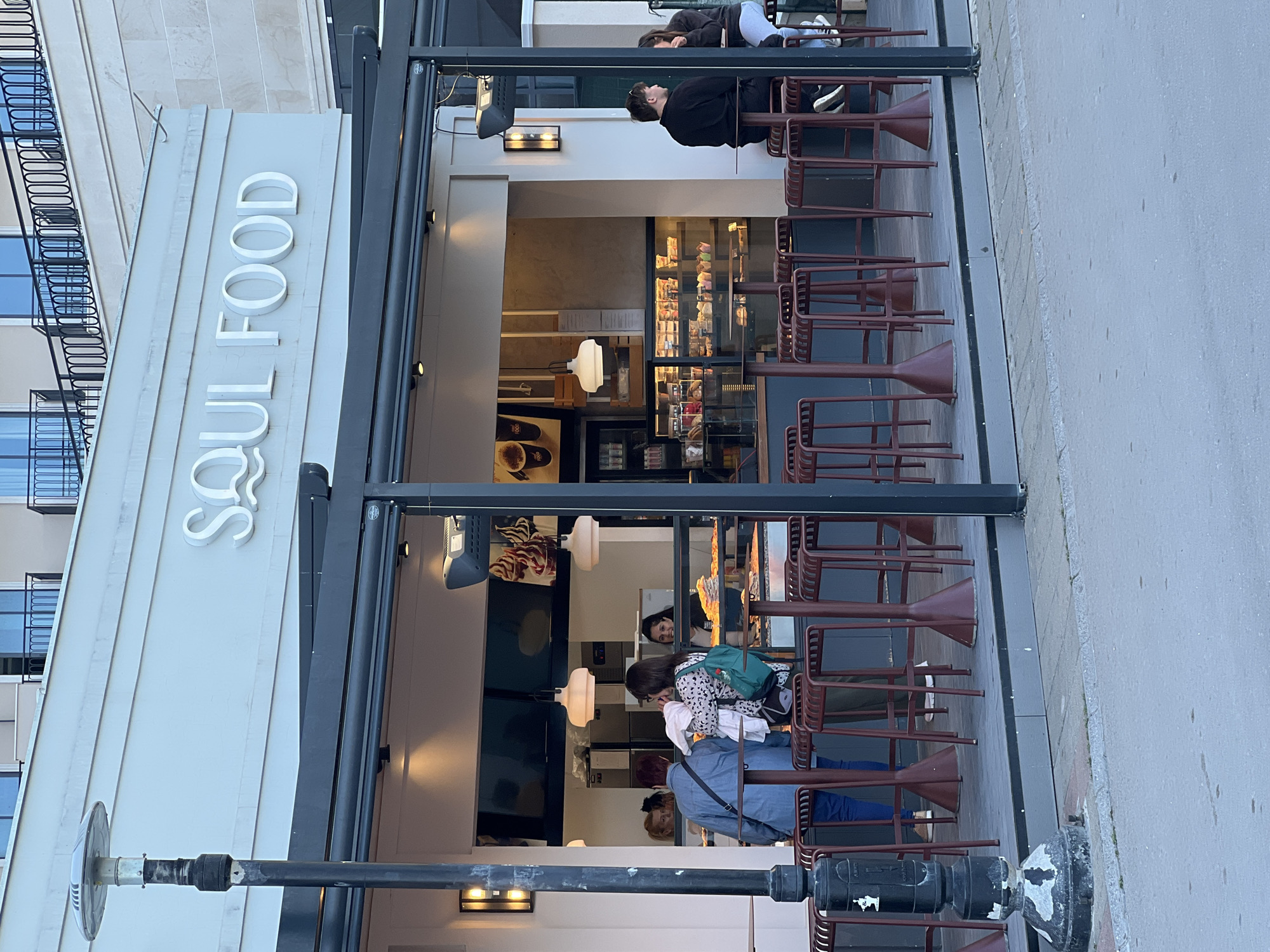 The image depicts the exterior of "Soul Food," a restaurant in Budva, Montenegro. The restaurant has a gray facade with a white sign reading "Soul Food" in capital letters. The entrance is framed by a black awning structure, with people seated at high-top tables with maroon chairs underneath. One person is leaning over the table, while others are standing or seated, possibly interacting. Inside, a display case is visible, filled with food items. The street is gray, and a lamp post is positioned in front of the restaurant. A building with windows is visible in the background.