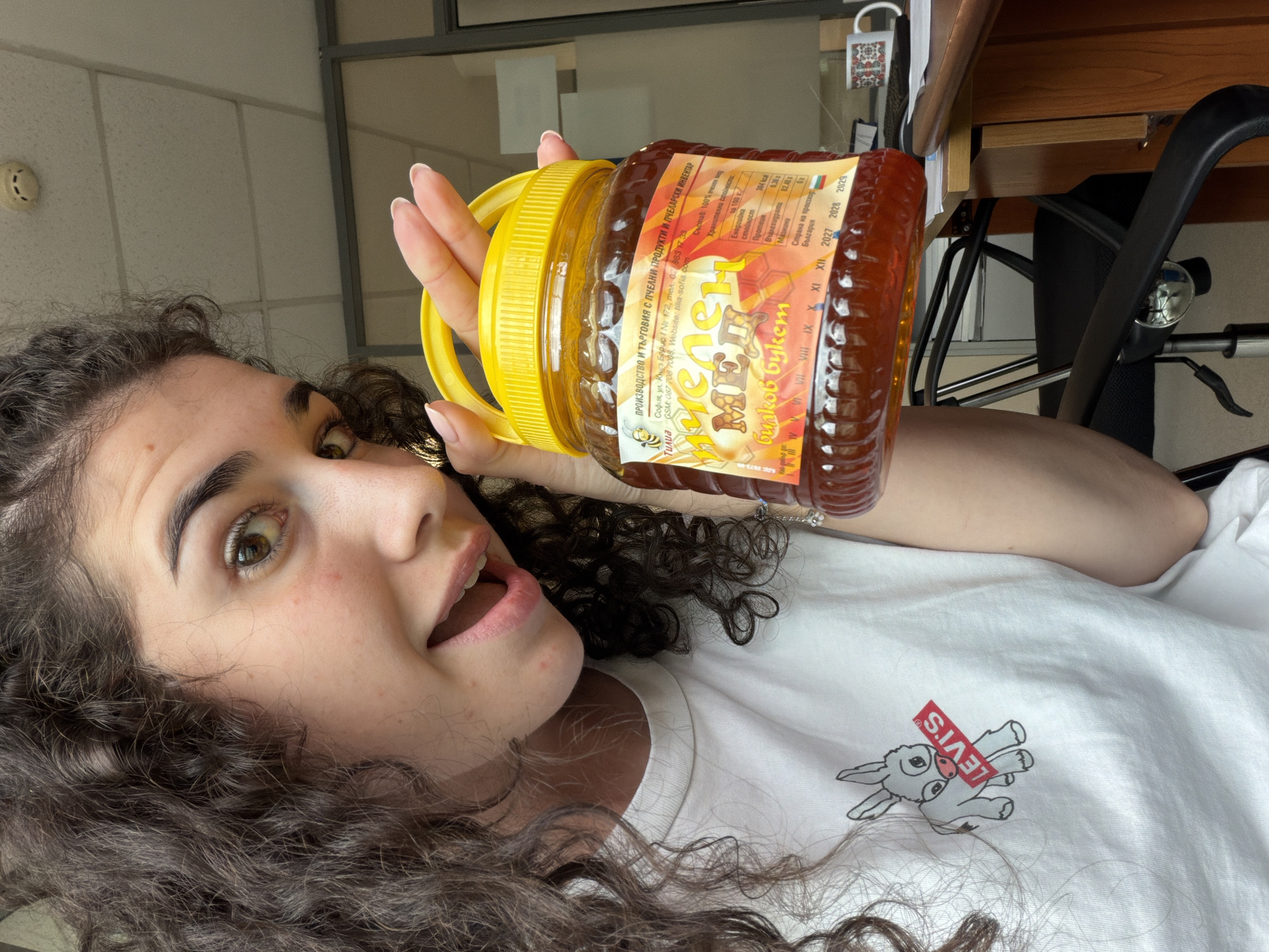 A young woman with dark curly hair is holding a large plastic jar of honey. She is wearing a white t-shirt with a cartoon donkey wearing a red Levi's tag. The honey jar has a yellow lid and a label with text in Cyrillic. The label indicates the honey is from "Тилия" (Tilia), a company located in Sofia, Bulgaria, at "София, ул. Княз Борис I № 172, тел. 087 9883 17, 33." The background shows an office environment with a desk, chair, computer monitor, and a mug with a folk design. The woman is looking at the camera with her mouth open in a surprised expression.