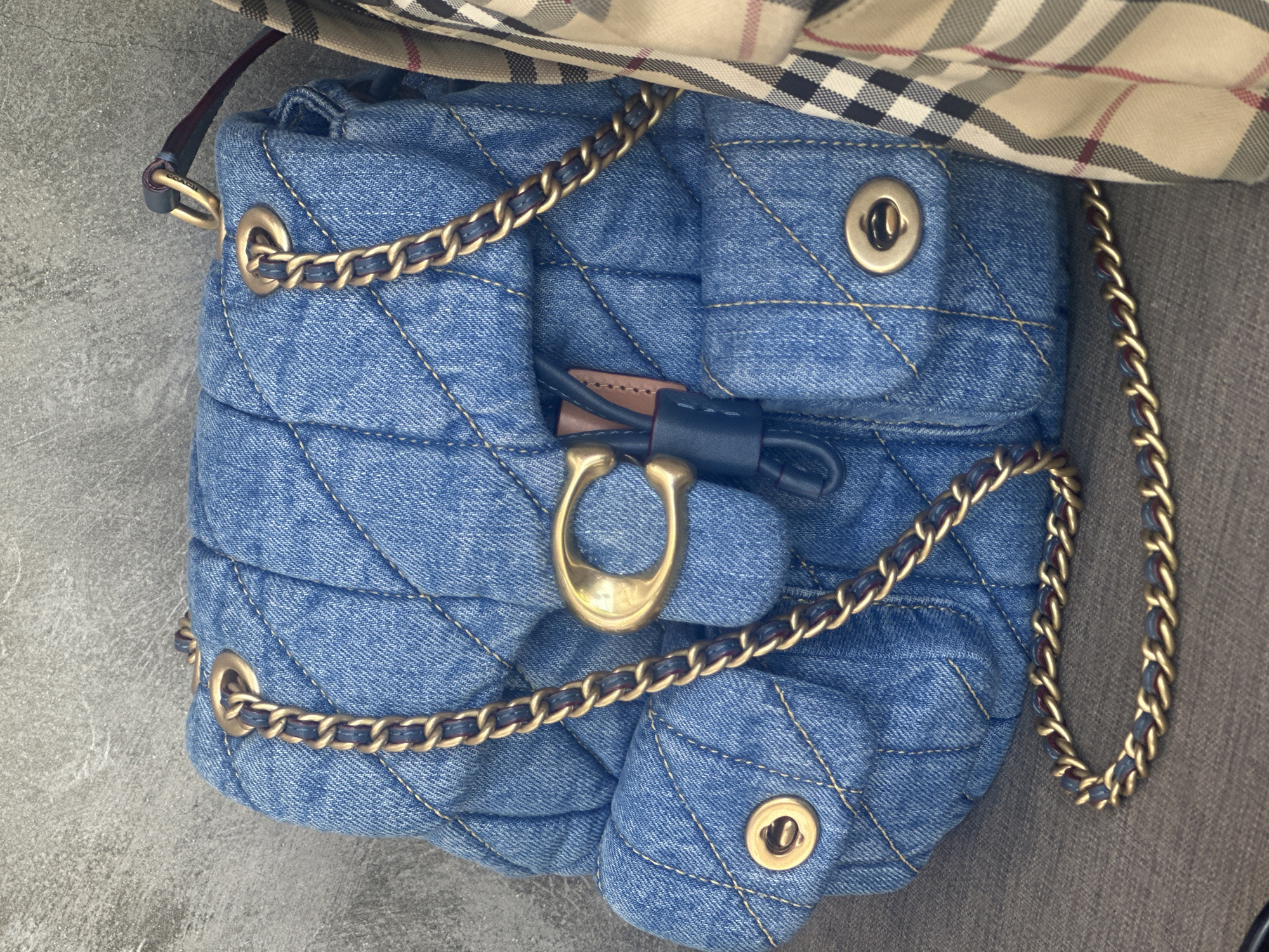 A quilted denim backpack with gold chain straps is displayed against a textured gray wall. The backpack features two front pockets with gold hardware and a flap closure with a gold "C" logo. A tan and plaid scarf is draped over the right side of the backpack. The backpack is resting on a textured gray surface.
