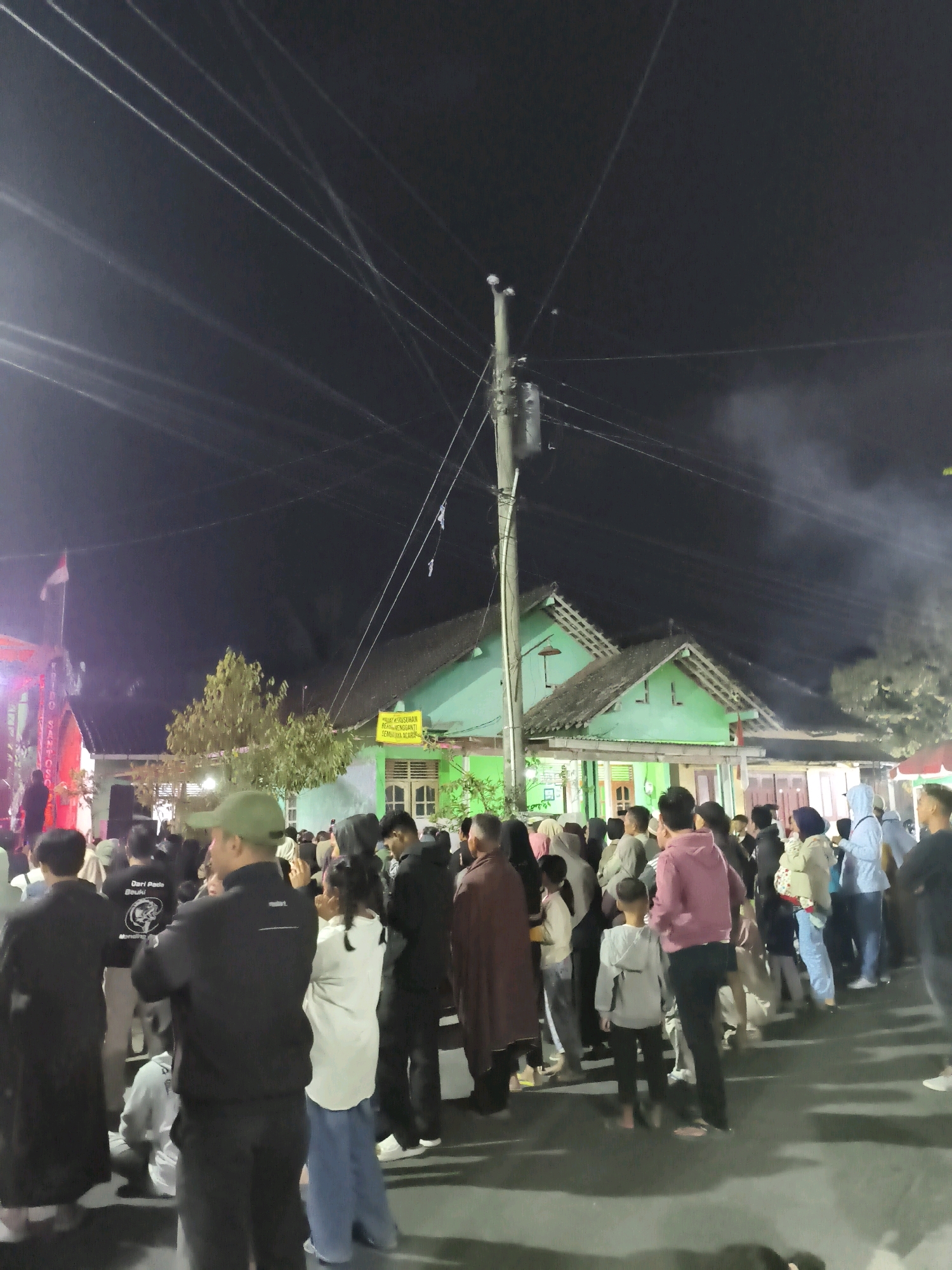 A nighttime, medium shot captures a crowd of people gathered on a street in Kembaran, Indonesia. The scene is illuminated by streetlights and the glow from a building with light green walls and a dark roof. Overhead, a network of power lines crisscrosses the dark sky, with a utility pole visible in the center of the frame. Several individuals are facing away from the camera, engaged in conversation or observing something out of frame. In the foreground, a person wearing a black jacket and a green baseball cap is partially visible, looking to the right. To the left of the frame, a red and white flag is flying. The ground is paved and appears wet, reflecting the surrounding lights. The overall impression is of a community gathering or event taking place after dark.