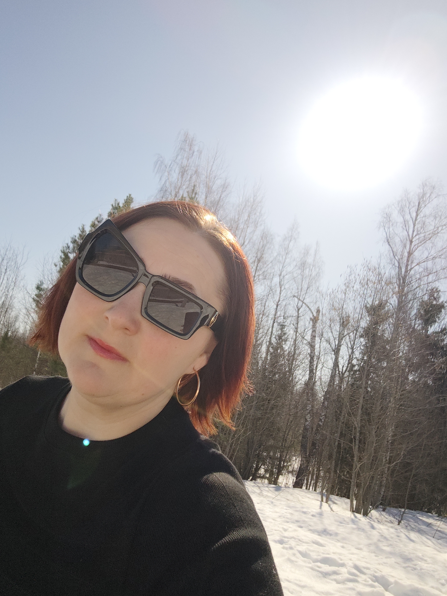 A woman with short red hair and oversized black sunglasses is taking a selfie outdoors on a sunny day. She is wearing a black top and gold hoop earrings. The background shows trees and snow, with the sun shining brightly overhead. The location is Марковский, Russia, based on the implied setting and reference information.