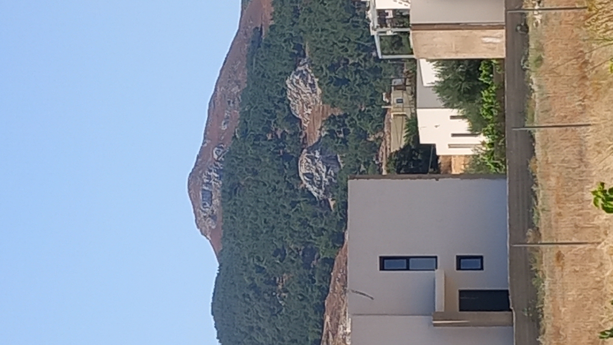 The image shows a landscape with a mountain covered in trees in the background and modern buildings in the foreground. A rocky outcrop is visible on the mountainside. The sky is clear and blue. The buildings are white with dark windows and doors. There is dry grass in the foreground, and a fence runs across the bottom of the image. The location is stated as Άγιος Νεκτάριος, Greece.