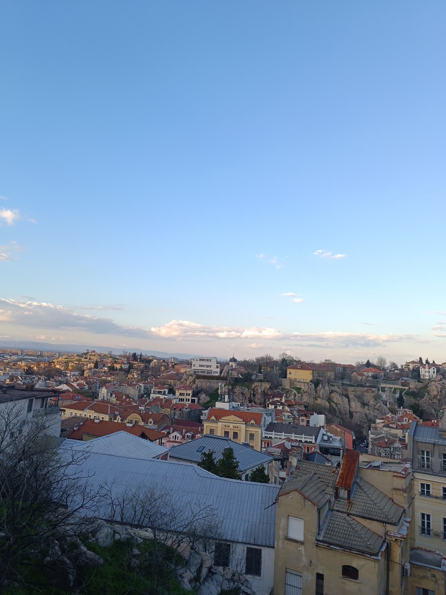 The image presents a high-angle view of Plovdiv, Bulgaria, under a blue sky with scattered clouds. In the foreground, there are rooftops of buildings constructed in a variety of styles and colors, some with reddish-orange tiles, others with gray metal sheets. Some of the buildings are old and appear weathered, while others look more modern. Bare tree branches are visible in the lower left corner, suggesting it might be late autumn or early spring.
In the middle ground, there are rock formations amongst the buildings, indicating the unique topography of Plovdiv. Some buildings are built on or around these rocky hills. In the distance, there's an indistinct skyline of hills or mountains on the horizon. The overall impression is of a densely built urban environment interspersed with natural geological features.