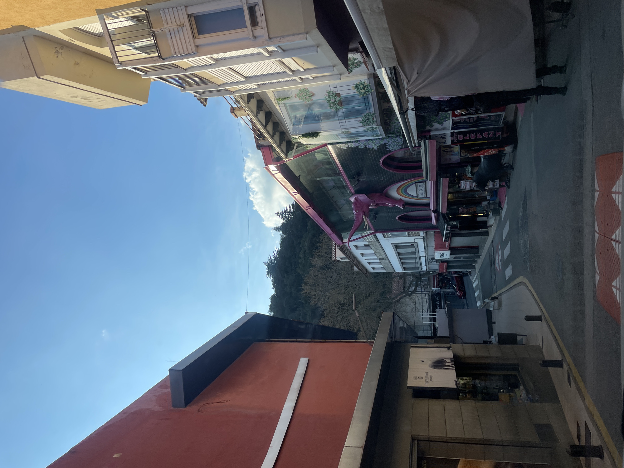 The image depicts a narrow urban street in La Jonquera, Spain, viewed from a slightly elevated, forward-facing perspective on a clear day.
On the left side of the street, a large building with a uniformly painted reddish-orange facade dominates the foreground. A dark grey horizontal structure extends from the upper part of this building. Below it, a commercial storefront features dark glass and a prominent white sign displaying the text "TERRITUMONTA" and "PERFUMES" or similar, alongside a silhouette of a quadruped animal. The sidewalk adjacent to this building is paved with light grey slabs and delineated by several dark grey bollards. Further down the street on this side, a red vehicle is partially visible parked near another building.
The right side of the street features a row of multi-story buildings with varied architectural styles. The initial building on the right has a light beige facade with multiple balconies, one of which is equipped with white slatted shutters. Further along, a distinctive dark-colored building features a large pink elephant sculpture with tusks emerging horizontally from its upper facade. Below the elephant, arched openings with pink trim are present, and a rainbow motif is painted above one arch. A sign reading "LA K'VE NEGRE" is visible near the elephant. Adjacent to this, another section of a building displays a mural depicting window boxes with flowers. Multiple tan-colored awnings extend over the sidewalk from these storefronts. A black bull sculpture stands on the sidewalk near one of these awnings. Below the awnings, various commercial signs are visible, including a red vertical sign with "ELEFANT" in white lettering. One individual, wearing dark clothing, is partially visible from the knees down, standing on the sidewalk under an awning on the far right.
The asphalt street surface features white pedestrian crossing stripes and a white painted yield symbol. A dark circular manhole cover is present in the street. In the background, beyond the buildings, a densely forested hill or mountain covered with dark green trees rises against the horizon. The sky is predominantly clear light blue with a few scattered white cumulus clouds. Direct sunlight illuminates the scene, casting shadows towards the right side of the image.