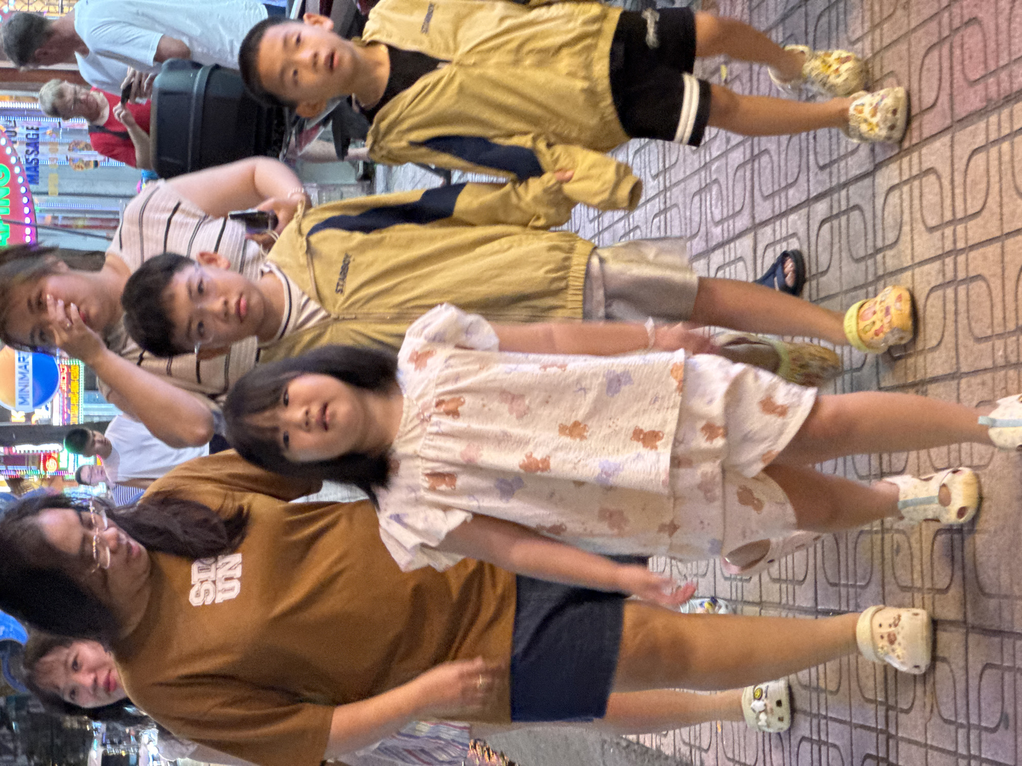 In Nha Trang, Vietnam, four children and two adults are walking on a tiled sidewalk at night. The group is comprised of two young girls, one in a light-colored pajamas with teddy bear prints, and a toddler in similar attire. They are accompanied by two young boys wearing yellow jackets over black t-shirts and shorts. Two adults, one wearing a brown t-shirt with the words "STUN" printed on it, and another woman in a striped shirt, walk with the children. In the background, neon lights from shops illuminate the street. The group appears to be walking towards the viewer, with the children in the front and the adults behind them.