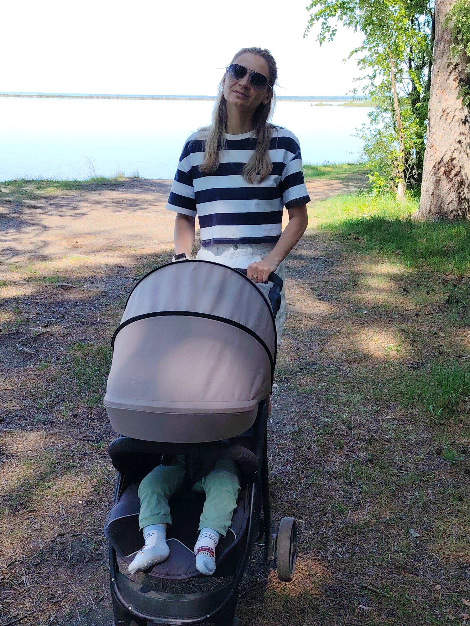 The image shows a woman pushing a baby stroller along a path. The woman is fair-skinned with blonde hair, wearing sunglasses, a striped t-shirt, and white shorts. The baby stroller is a dark color with a light-colored canopy. Inside the stroller, the baby is wearing green pants and white socks with a red design. The path is dirt and pine needles with green grass on either side. In the background, there is a body of water and trees. The location is outdoors in a natural area, likely in Chaikovsky, Russia, as requested.