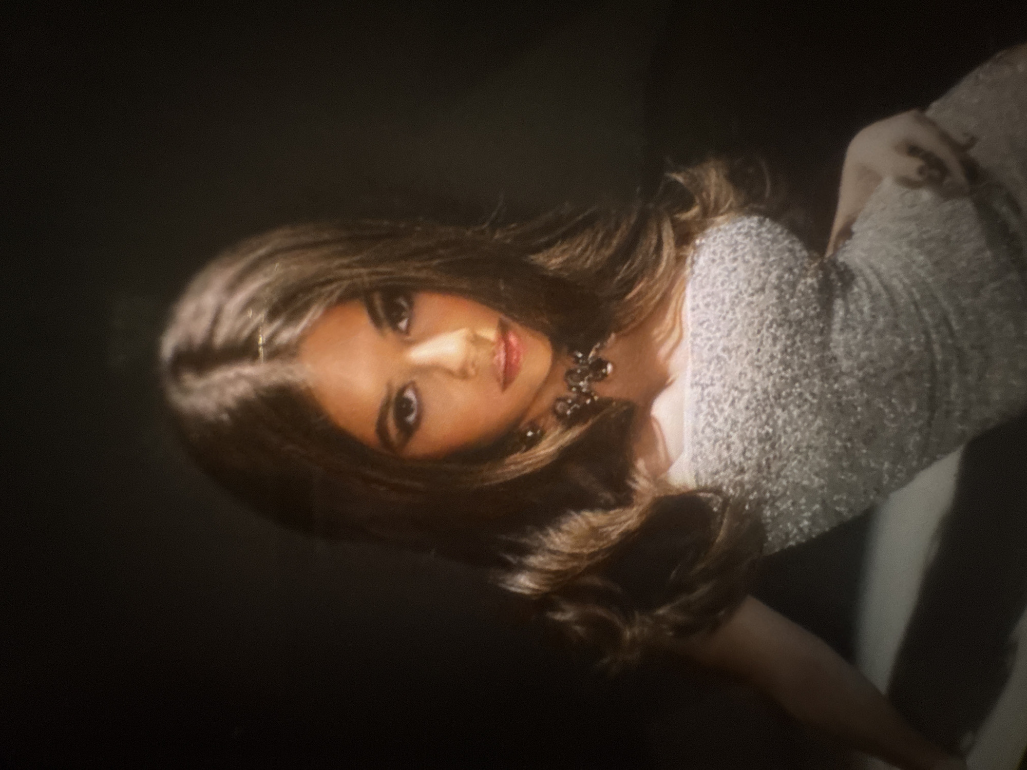 A medium close-up portrait features a woman with long, dark brown hair styled in soft waves, parted down the middle. She is wearing a light-colored, possibly sequined or beaded, strapless dress that shimmers. A prominent dark necklace, possibly beaded or jeweled, adorns her neck. She has a full face of makeup, with dark, defined eyes and reddish-pink lips slightly parted. The woman looks directly at the viewer with an intense gaze. The background is predominantly dark, suggesting a studio setting or low-light environment, with some lighter shadows or reflections visible towards the right side. The overall lighting highlights her face and upper body. There is no visible text.