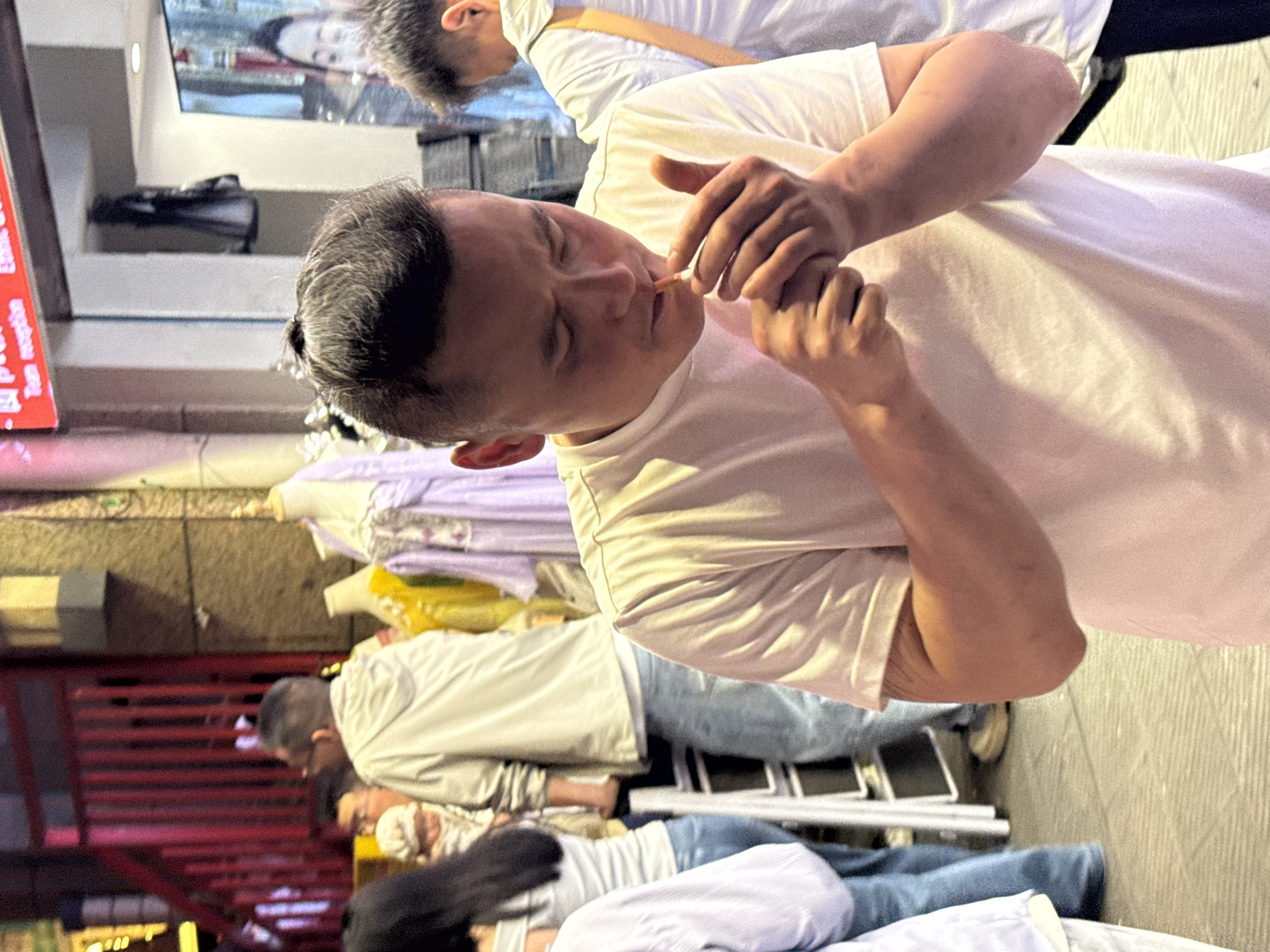 A man in a white t-shirt is lighting a cigarette with a match. He is standing outdoors, with other people standing or lying down in the background. The location is indicated as Yuzhong District, China, based on the photograph's origin.