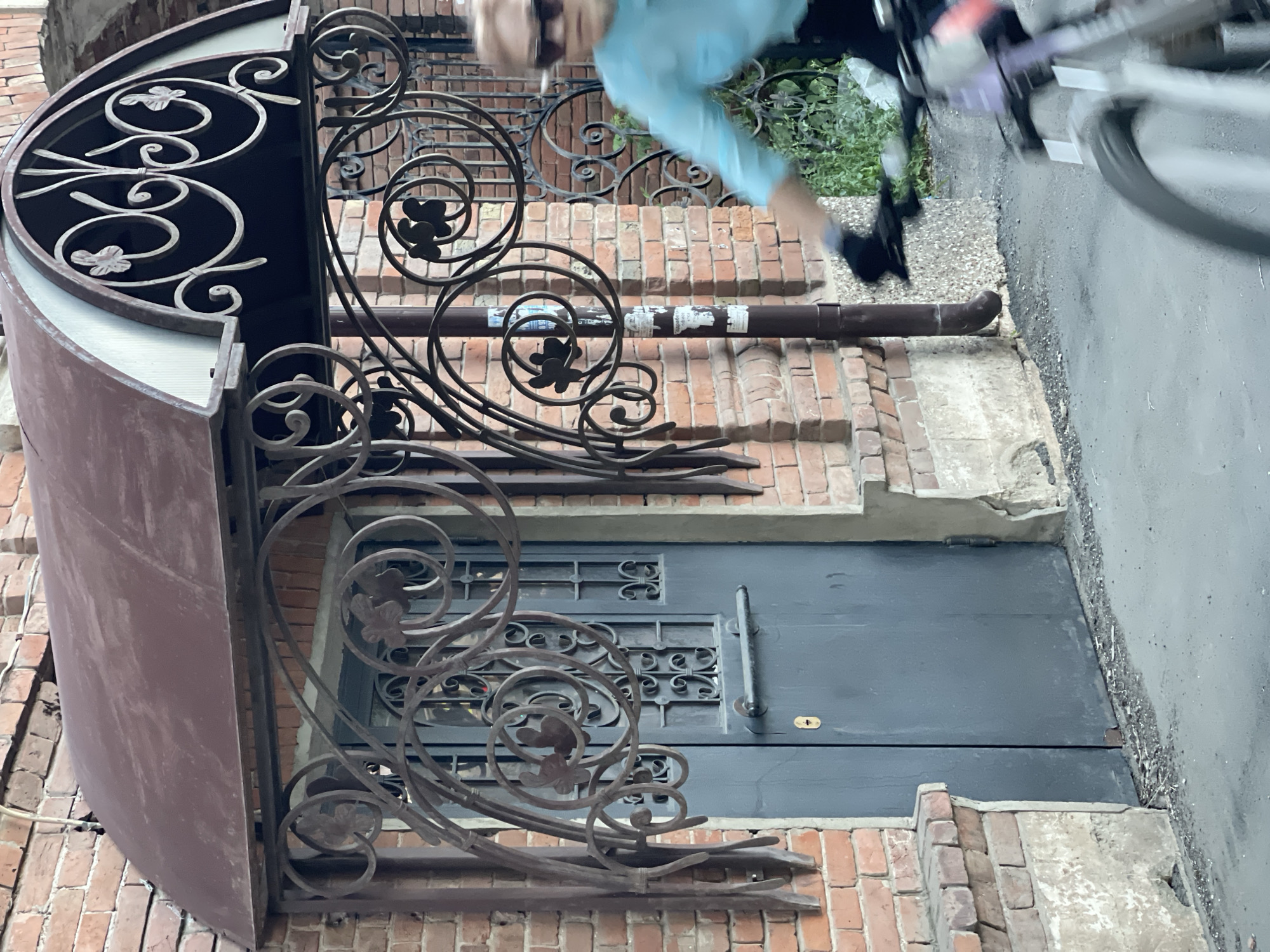 A woman wearing sunglasses and a blue shirt is riding a bicycle past a brick building with a dark gray door. The building has an ornate metal awning over the door, decorated with floral and scrollwork motifs. The street in front of the building is paved and has some debris along the edge. Green foliage is visible to the right of the building. The scene appears to be in an urban setting, possibly in Orenburg, Russia, given the context.