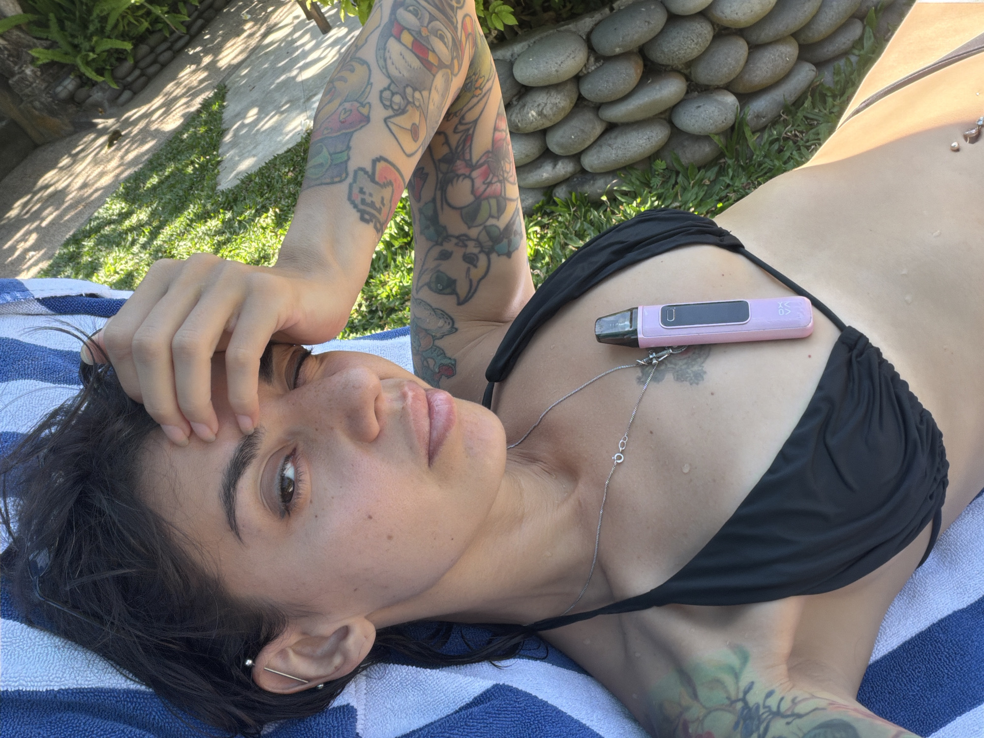 A person lies on a blue and white striped towel outdoors, their head resting on their arm. They are wearing a black bikini top and have multiple tattoos on their arms and chest. A pink vaping device is placed on their chest. The background shows green grass, a concrete path with dappled sunlight, and a wall of smooth, gray stones. The location is identified as Blahbatuh, Indonesia.
