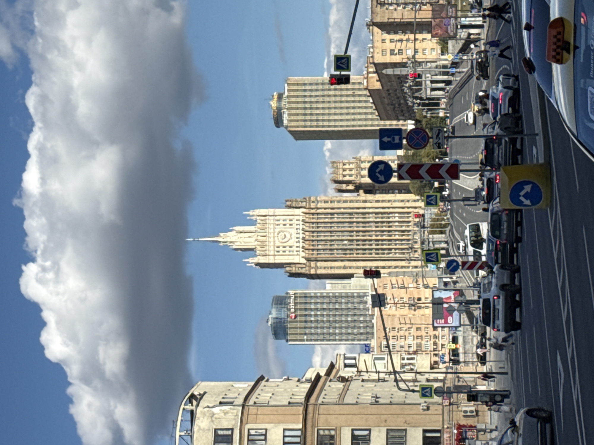 This image captures a street view in Moscow, Russia, focusing on the Stalinist skyscraper known as the Ministry of Foreign Affairs Building, a prominent landmark. The foreground features a multi-lane road with various vehicles, including cars, a white van, and a yellow taxi in the lower right. Pedestrians are visible crossing the street and walking on the sidewalk. Traffic lights and road signs, including directional arrows and a "no stopping" sign, are present. The middle ground displays a cluster of buildings, including a modern glass-fronted tower with the "azimut" logo. The background is dominated by the imposing Ministry of Foreign Affairs Building, characterized by its distinctive architecture, spires, and clock face. The sky above is blue with large, white clouds. The overall scene depicts a busy urban environment in daylight.