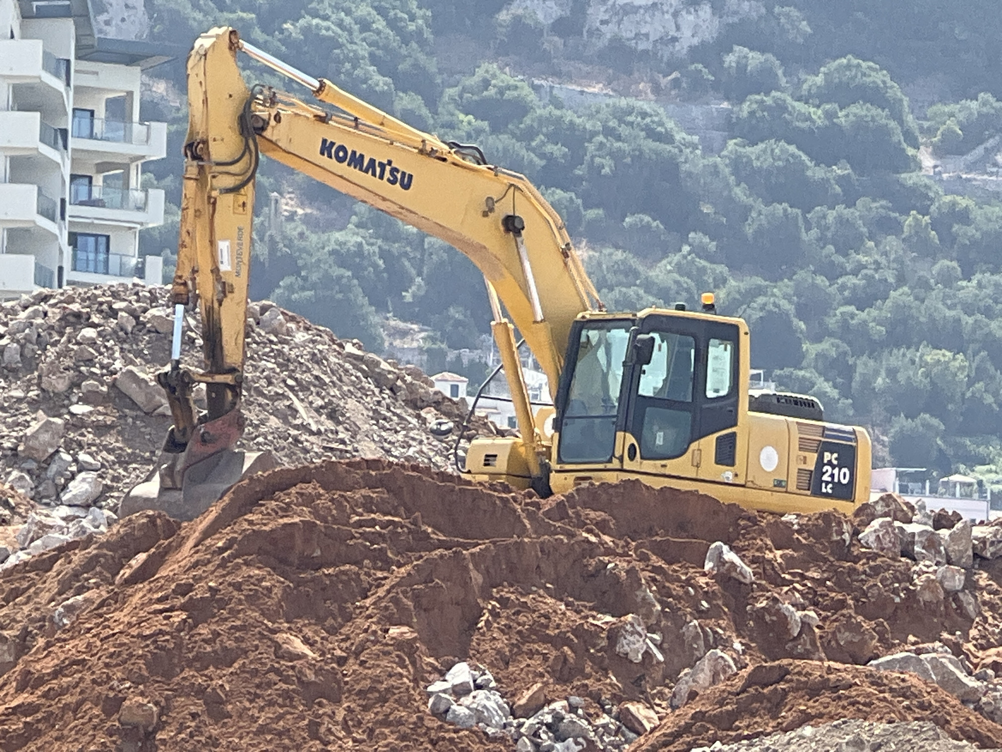 A Komatsu PC 210 LC excavator is actively engaged in demolition or construction work on a site in Gibraltar, Gibraltar. The excavator is positioned on a large mound of reddish-brown soil and debris, with its boom extended and bucket resting on the pile. In the background, a modern white building with multiple balconies and blue-tinted windows is visible. Beyond the building, a lush green, tree-covered hillside characteristic of Gibraltar's terrain rises steeply. The sky above is overcast, creating a diffused light. The scene suggests ongoing development or site preparation within the urban and natural landscape of Gibraltar.