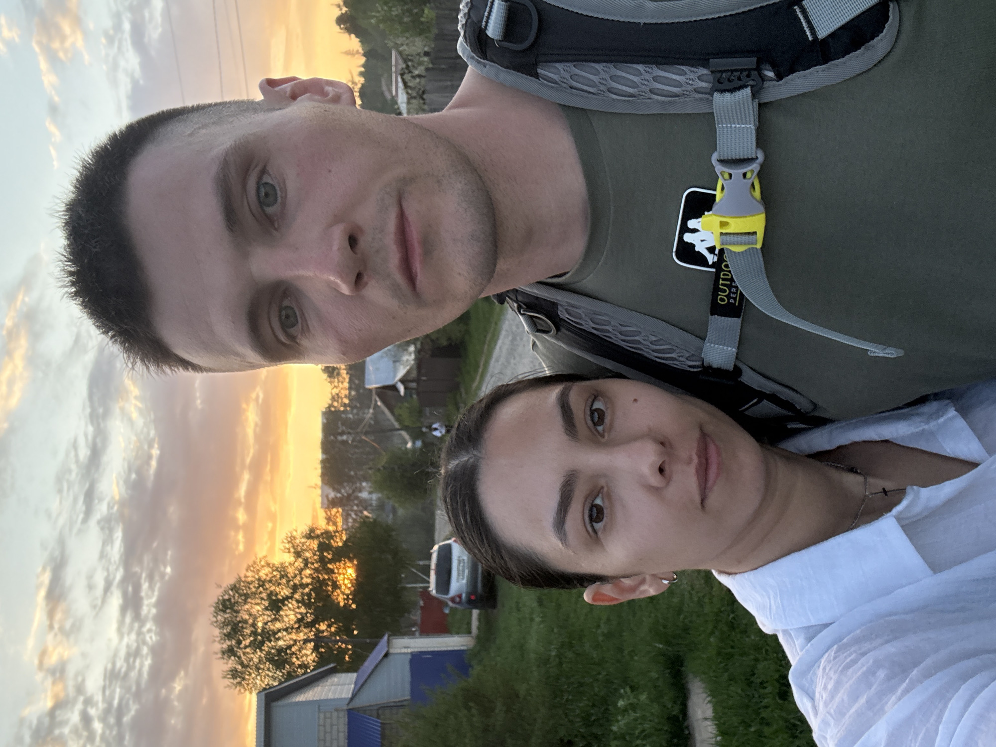 A selfie-style photograph shows two people outdoors at what appears to be dusk or sunset. A man in an olive green t-shirt and wearing a backpack is in the foreground. To his right and slightly below him is a woman wearing a white blouse. Behind them is a residential area with houses, trees, and a sky with orange and white clouds, typical of a sunset. A white van is parked near one of the houses. The location might be Chaikovsky, Russia, given the appearance of the buildings and the overall environment. The image suggests an informal setting, possibly taken during a walk or outing.