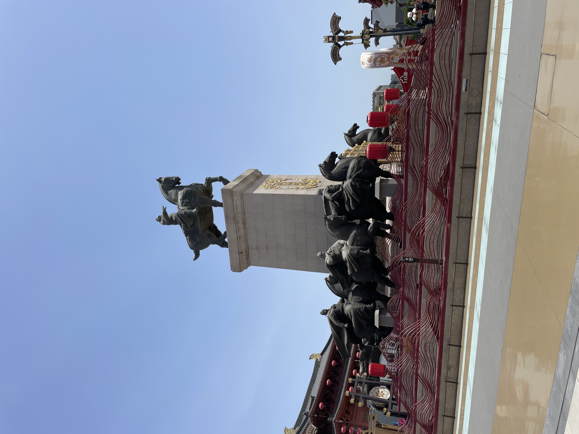 The image depicts a monument in Yanta District, China. A bronze statue of a man on horseback is perched atop a tall, rectangular stone pedestal. Below, a series of bronze sculptures of figures and horses line the structure. Red lanterns and a decorative red railing adorn the scene. A traditional-style building with a dark roof and ornate details sits at the base. The sky is a clear, bright blue.