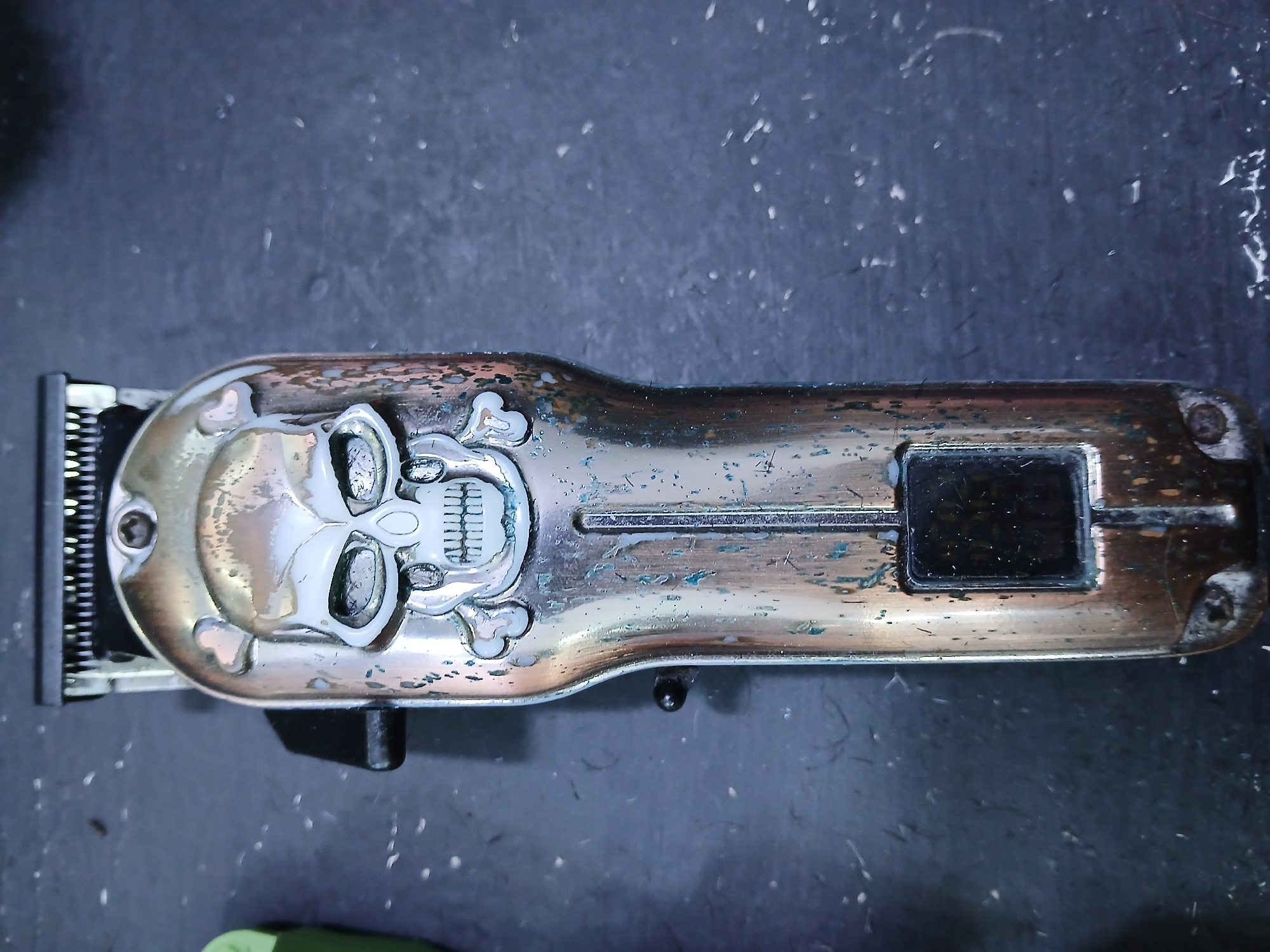 The image shows a hair clipper with a skull design lying on a dark, possibly metallic surface. The clipper has a metallic finish that appears worn and weathered with patches of discoloration. The skull design is prominently featured on the clipper's head. The clipper's blades are visible on the left side of the image. There is no evidence to suggest a location in the city of Juan Griego, Venezuela.