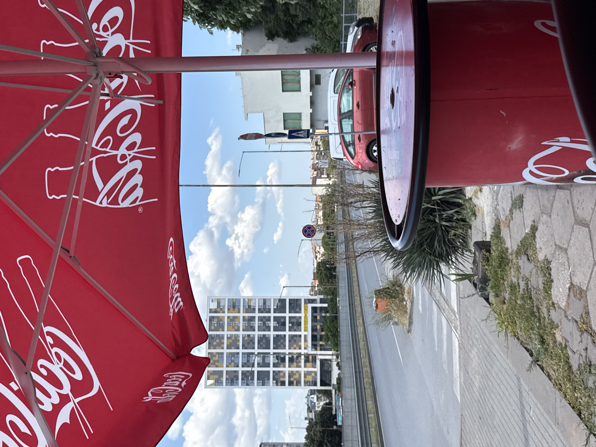 The image depicts a street scene in Varna, Bulgaria, on a cloudy day. In the foreground, a red Coca-Cola branded umbrella covers a table and its base. Beyond the umbrella, a two-lane road runs parallel to a sidewalk. A white minivan and a red hatchback are visible on the road. In the middle ground, a tall, modern building with a grid of windows, some of which are yellow and grey, dominates the skyline. Behind it, other buildings and trees are visible. The sky is blue with scattered white clouds.