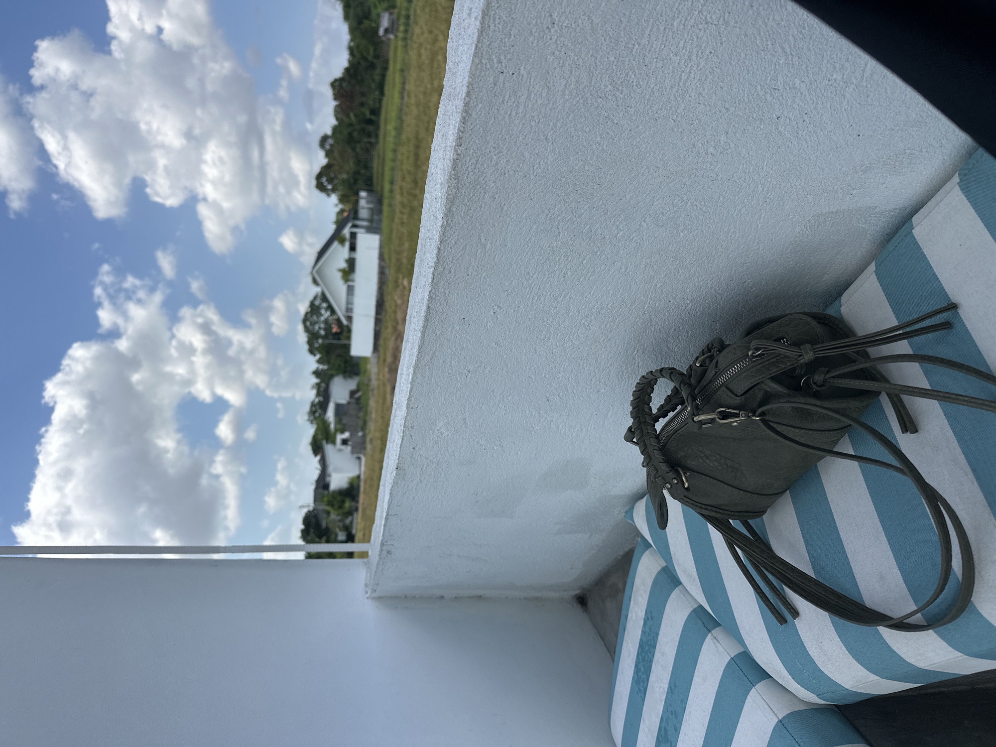 An outdoor scene in North Kuta, Indonesia, captures a glimpse of a residential area under a bright, cloudy sky. In the foreground, two blue and white striped cushions rest on what appears to be a seating area. A dark green, cylindrical handbag with braided handles and fringe details is placed on the cushions. The background reveals a landscape with scattered modern white houses, lush green trees, and a field of dry grass or crops. The architecture suggests a tropical or subtropical location. The scene is static, with no people or active engagements depicted.