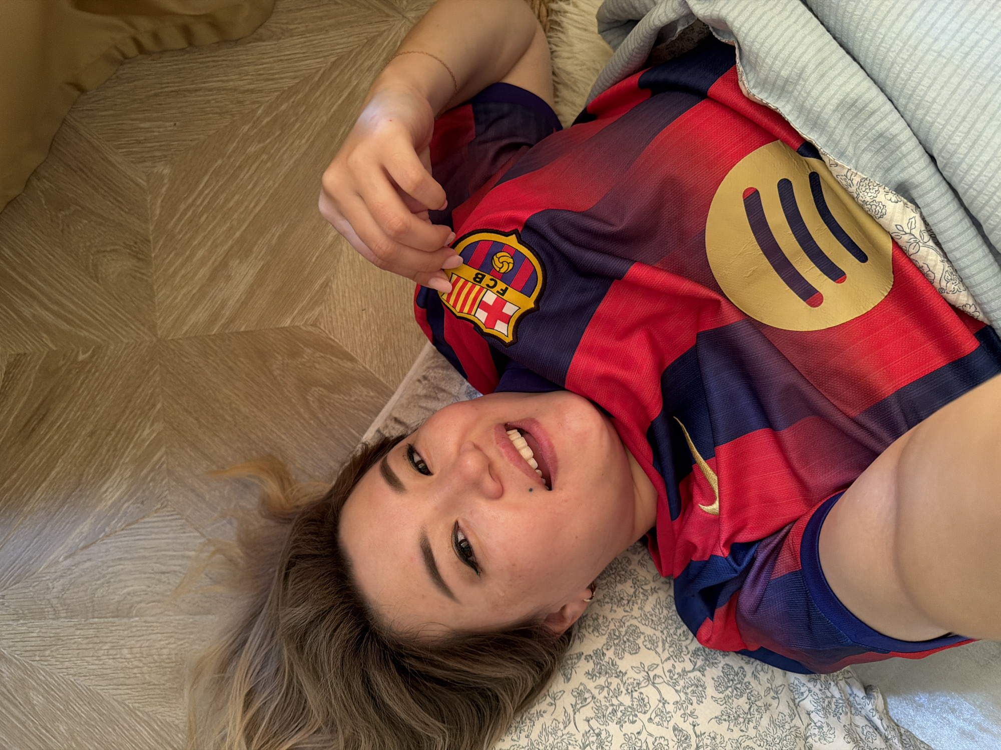 A woman is lying on a bed in Astana, Kazakhstan, wearing a red and blue striped FC Barcelona jersey. The jersey has a Spotify logo on the left chest and the FC Barcelona crest on the right chest. The woman is looking at the camera and smiling. Her hair is blonde and her makeup includes winged eyeliner and lip gloss. A patterned blanket is visible to her left, and a wooden floor is to her right.