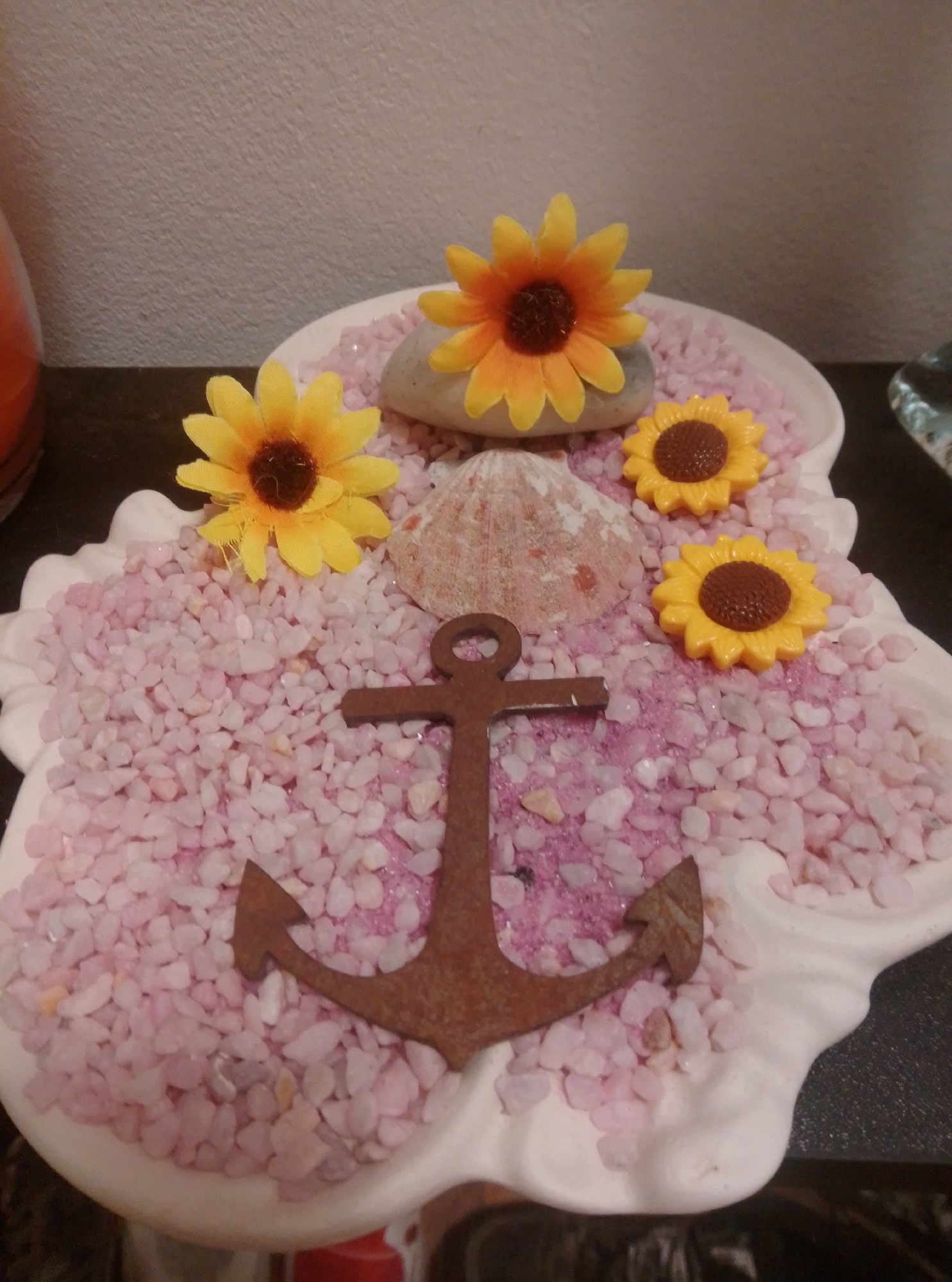 A decorative display is set on a dark surface, likely a table, in Wedel, Germany. The display features a white, scalloped dish filled with small pink and purple gravel. Several decorative elements are placed within the gravel. On the left, a yellow artificial sunflower with a brown center rests on a light grey rock. To its right, a beige seashell is positioned. Further to the right, two smaller yellow artificial sunflowers with brown centers are placed. In the foreground, a rusty brown anchor ornament is positioned horizontally across the gravel. The background shows a textured wall and part of an orange object.