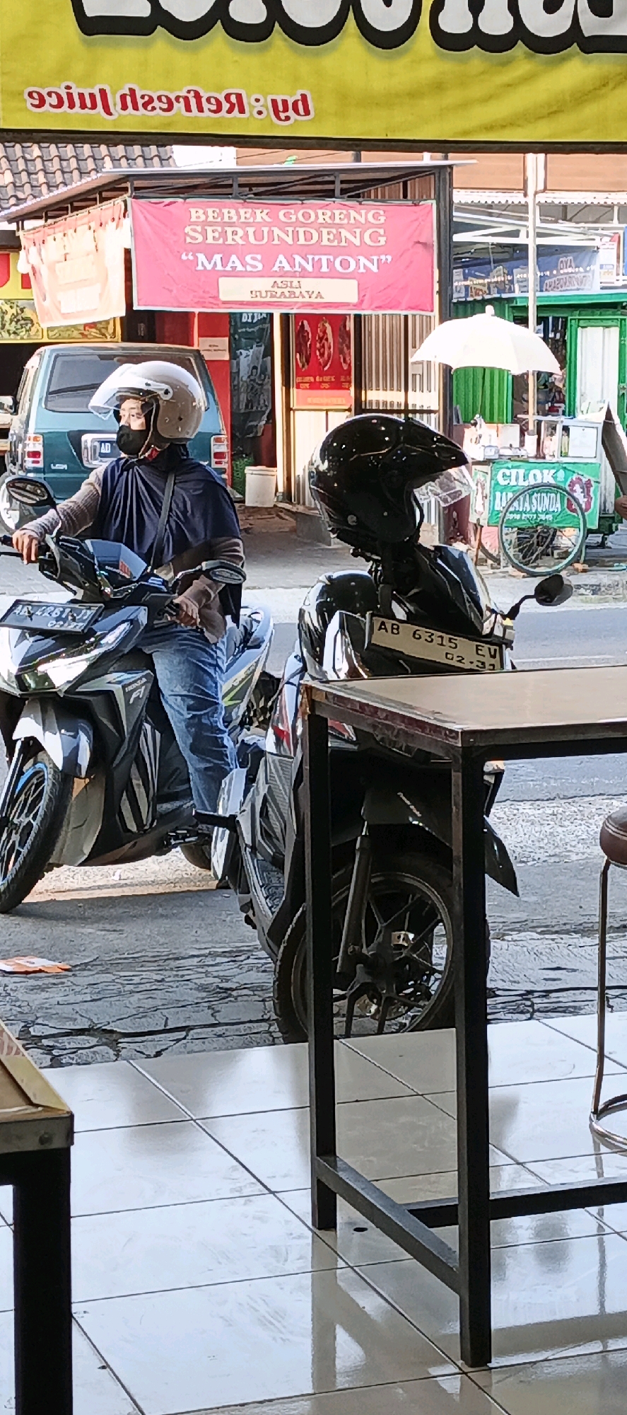 In Sidoarum, Indonesia, an outdoor street scene is depicted. In the foreground, a table with a metallic frame and a wooden top occupies the lower right portion of the frame, casting a shadow on the tiled floor. To the left of the table, a black motorcycle is parked, its front wheel and part of its body visible. A black helmet rests on the back of this motorcycle, obscuring its license plate which reads "AB 6315 EV".
Behind the table and motorcycle, a person wearing a white helmet and a dark blue cloak over a brown shirt and blue jeans is seated on another motorcycle. The person is looking to the left. Their motorcycle's license plate is partially visible and reads "AB 4261 JB".
Further back, a pink banner with white text hangs across the street. The banner reads "BEBEK GORENG SERUNDENG 'MAS ANTON'" and "ASLI SURABAYA". Behind the banner, a green vehicle is parked. In the background, a white umbrella is set up, and other structures and vehicles are visible. The ground appears to be paved with tiles in the foreground and asphalt or concrete further back. The overall lighting suggests it is daytime.