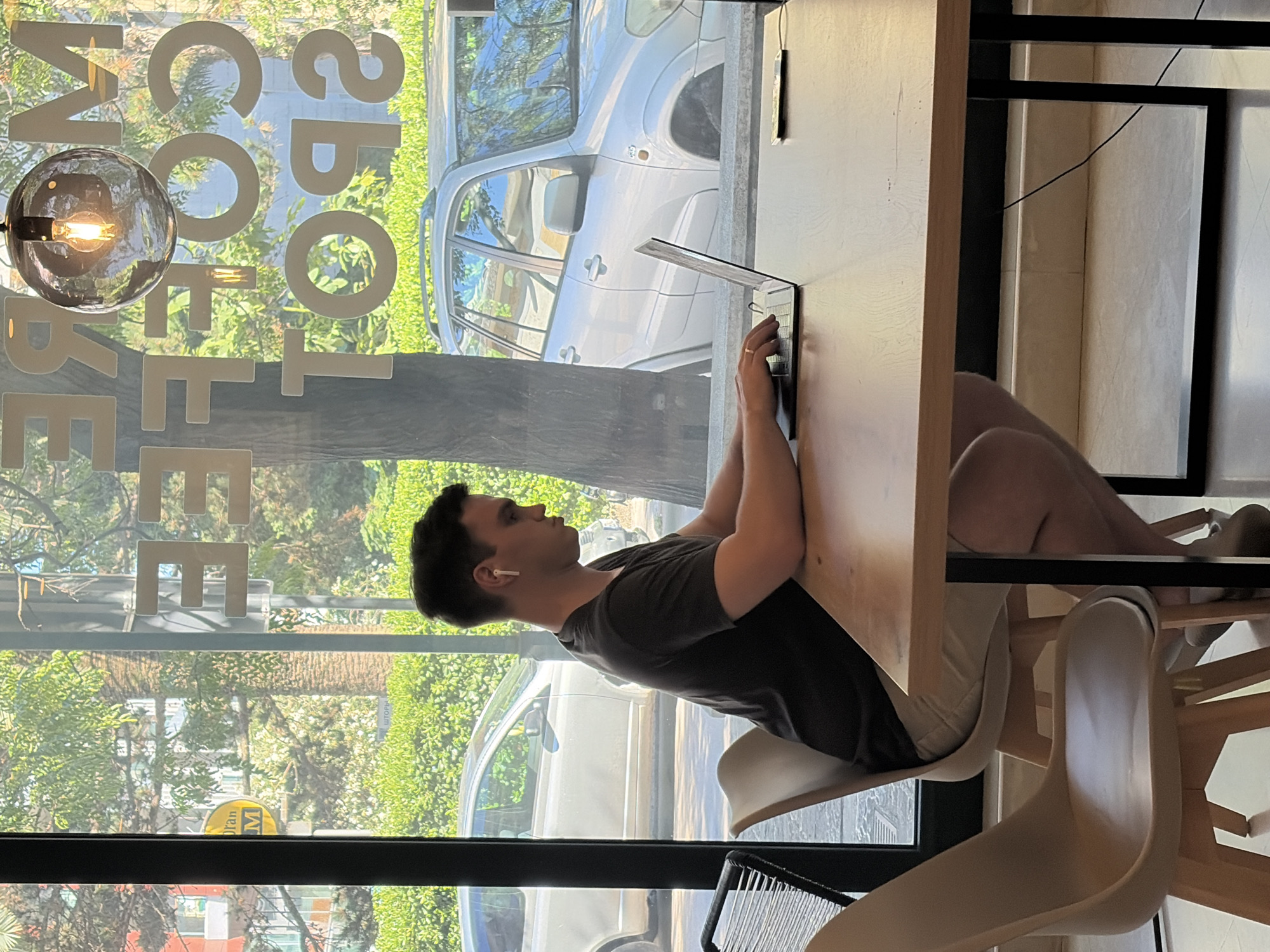 A young man wearing a dark t-shirt and light-colored shorts is sitting at a wooden table in a cafe, working on a laptop. He has earbuds in and appears focused on his screen. The cafe has large windows that look out onto a street with lush green trees and a silver SUV parked outside. There is a circular light fixture hanging from the ceiling, and text that reads "MORE COFFEE SPOT" is visible on the window. The floor is tiled. The cafe is likely in Budva, Montenegro, given the background scenery and the style of the cafe.