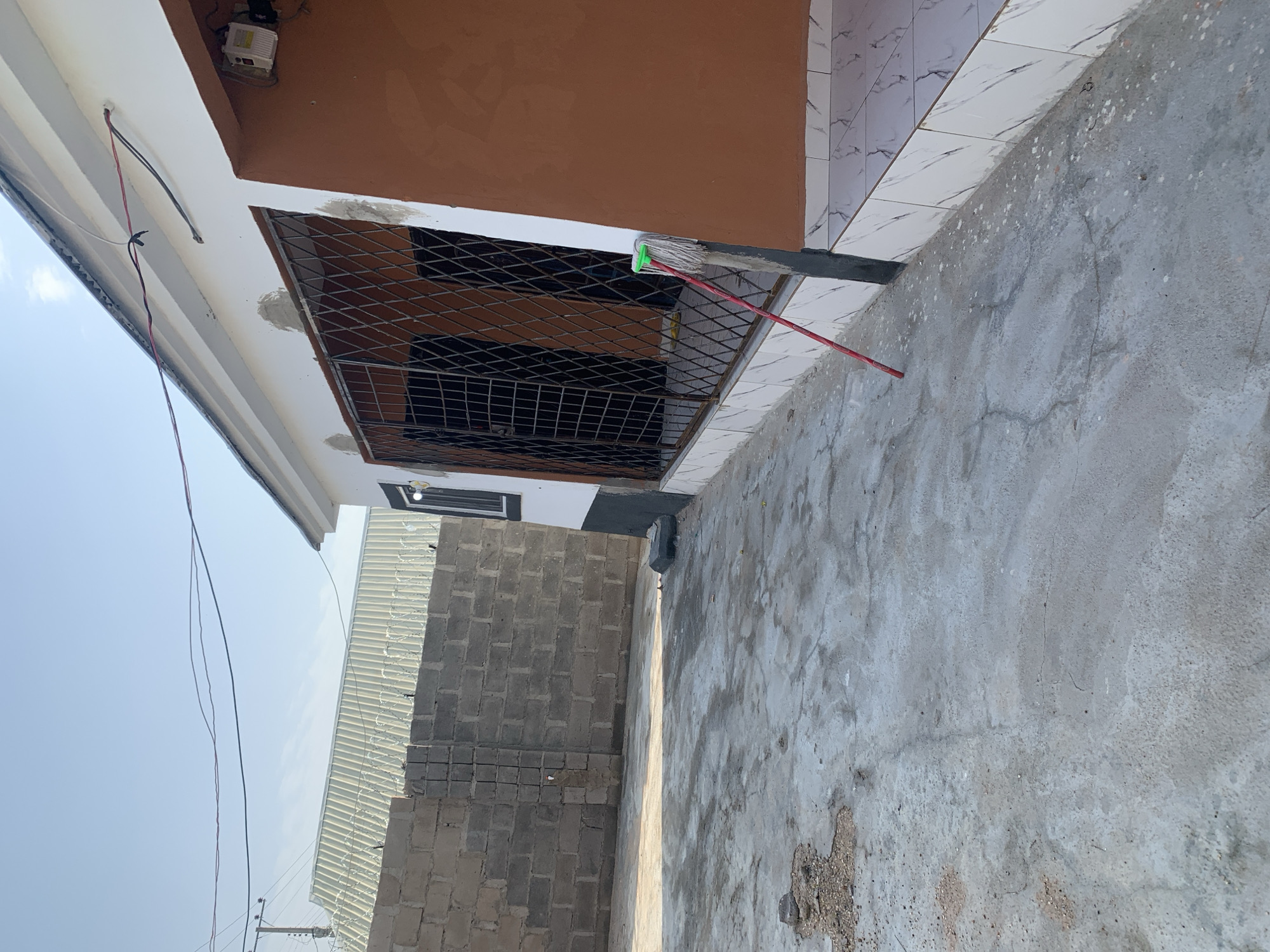 The image shows the exterior of a building with a concrete courtyard in front of it. The building has a white facade with an orange section and a metal security grate over a window. A mop with a green head and red handle is leaning against a vertical beam near the window. To the left of the building is a brick wall topped with barbed wire, behind which is a corrugated metal roof. The courtyard is cracked and shows signs of wear. The sky is clear and blue. The location is described as Unknown, Unknown.