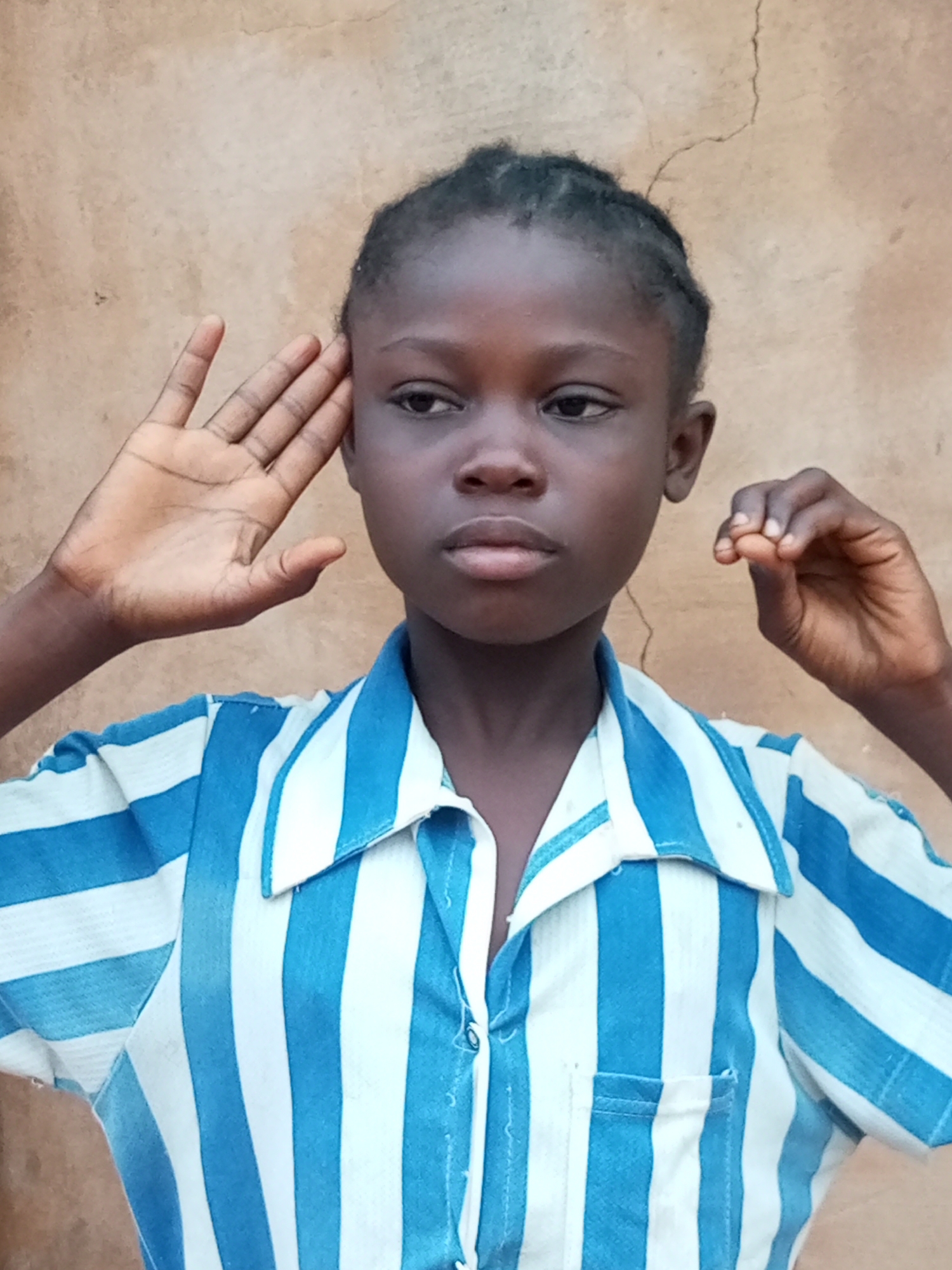 A medium shot captures a young Black person in what appears to be a rural or semi-rural setting in Tempure, Nigeria. The subject, likely a child or adolescent, is positioned against a textured, light brown wall that shows some signs of cracking. They are wearing a collared shirt with alternating vertical stripes of bright blue and white. Their left hand is raised, palm facing outwards, near their ear, as if listening or blocking sound. Their right hand is also raised, with fingers gently curled. The subject's gaze is directed slightly to the left and downwards, with a neutral expression. The lighting suggests it is daytime.