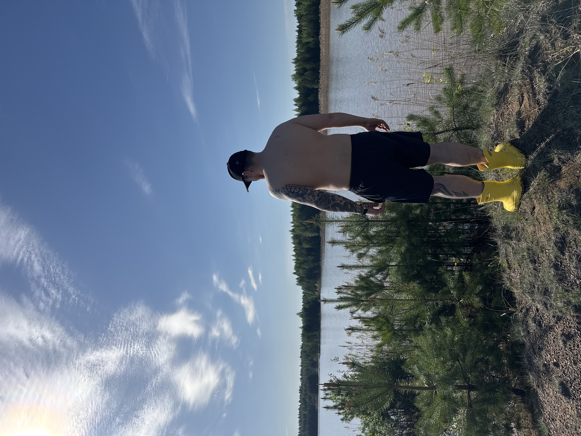 A man wearing a black baseball cap, black shorts, and yellow boots stands with his back to the viewer near a body of water, possibly a lake, in a forested area. The sky is blue with scattered clouds. The location is Noviy, Russia. The man has visible tattoos on his left arm. He stands on an embankment covered in grass and small trees. A line of trees is visible across the body of water.