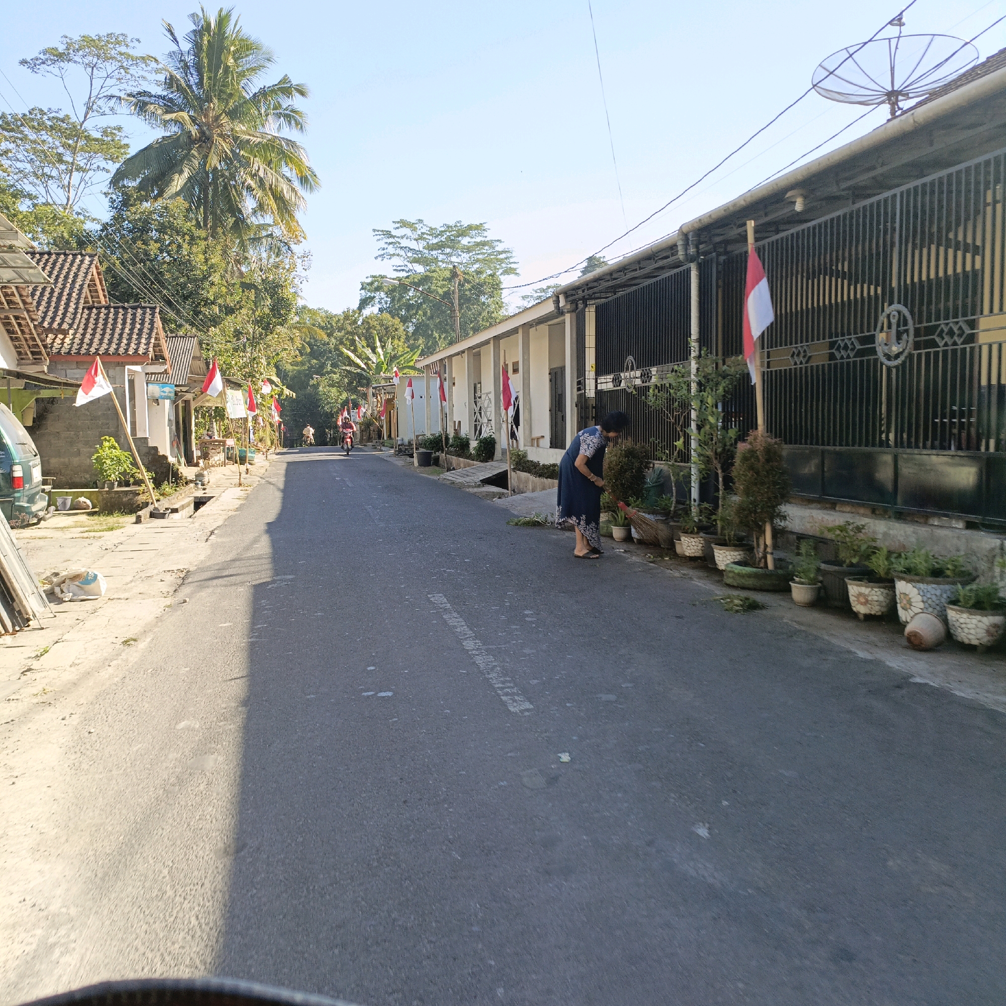 This image depicts a street scene in Salatiga, Indonesia, likely during a patriotic celebration given the prevalence of Indonesian flags. The street is paved with asphalt and bordered by residential buildings and a fence. A woman in a dark blue dress is sweeping the ground near potted plants lining the property. A person on a motorcycle can be seen further down the street. The weather appears clear and sunny, with a bright blue sky overhead. Palm trees and other lush greenery are visible in the background, suggesting a tropical or subtropical climate. The overall impression is of a quiet residential area undergoing some form of local observance.