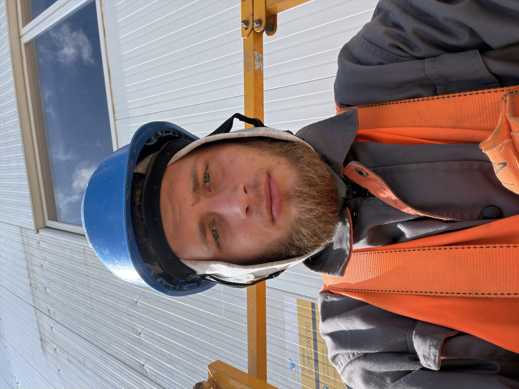 A man wearing a blue hard hat, a grey work jacket, and an orange safety vest is looking directly at the camera. The man has a beard and is standing in front of a white corrugated metal wall, with a window visible in the upper portion of the background. A yellow lift platform is partially visible on the left and right sides of the frame. There is text on a yellow sign partially visible on the left. This image was taken in the city of Stróża, Poland.