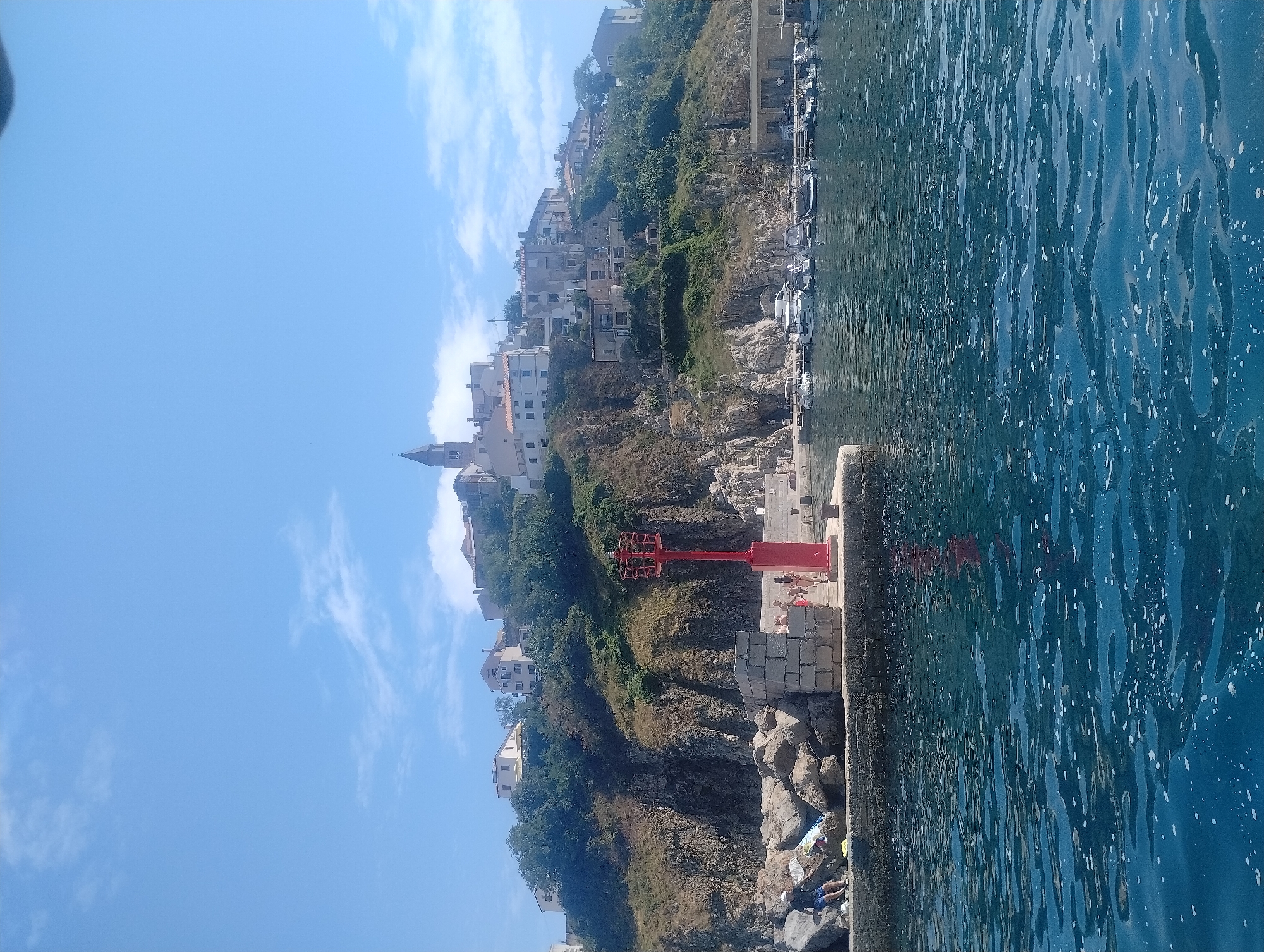 The image shows a body of water in front of a town built on a hill. On the left side of the image, there is a stone pier with large rocks and a red lighthouse. A person in blue shorts and a dark shirt is sitting on the rocks. Several other people are visible near the lighthouse and on the pier. The town on the hill features white buildings with red tile roofs, and a tall church steeple. The buildings are nestled among green trees and rugged cliffs. Boats are moored in the water in the background. The sky is blue with scattered white clouds. The location is Općina Vrbnik, Croatia.