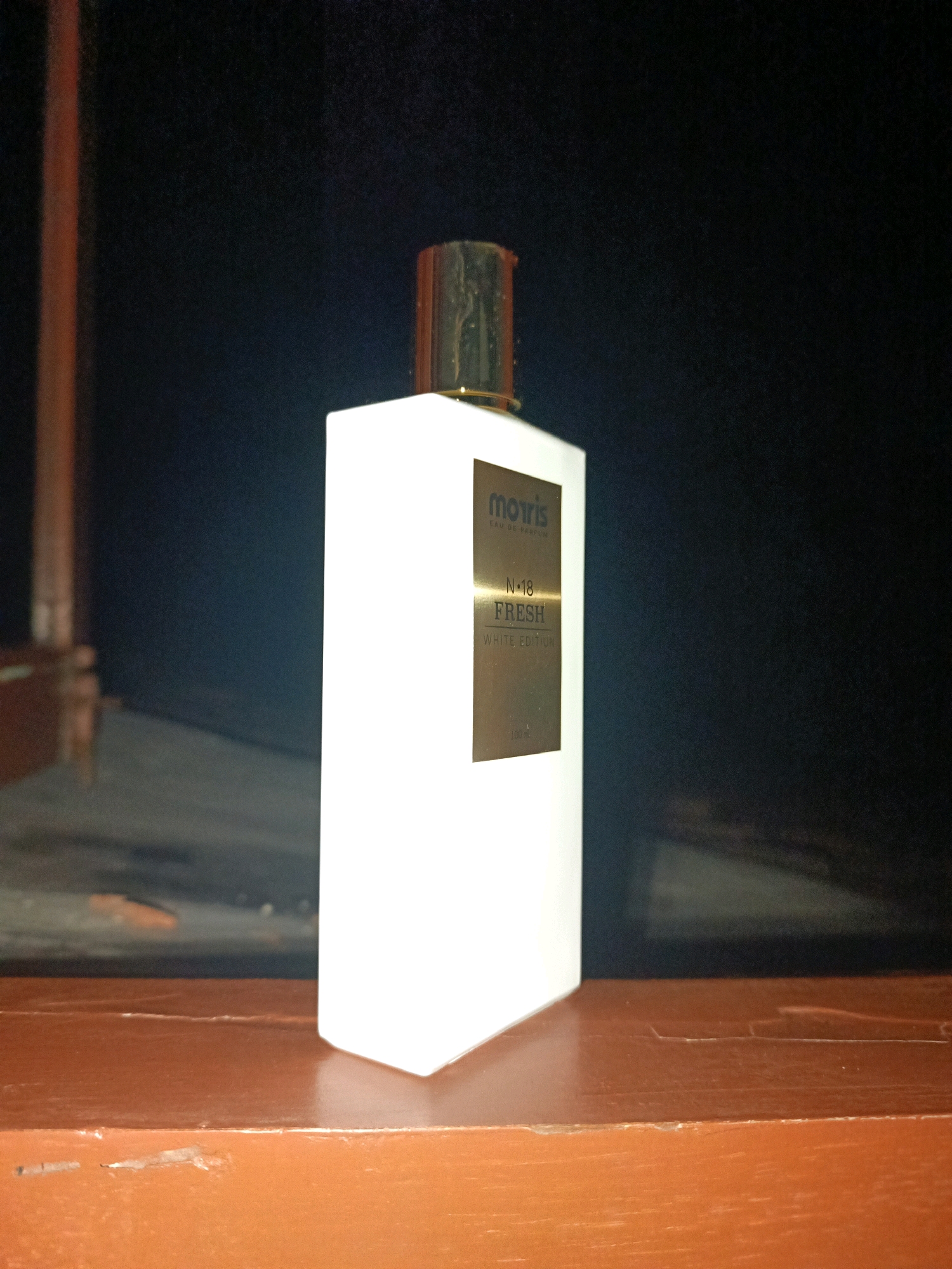 The image shows a white perfume bottle with a gold cap and a gold label. The label reads "morris EAU DE PARFUM N.18 FRESH WHITE EDITION 100 ml". The bottle is standing on a brown wooden surface. In the background, there is a dark, blurry area that appears to be a wall or a curtain, with some indistinct shapes that might be furniture. The lighting is dim, casting shadows and highlighting the bottle. The setting is not identifiable as being in Sragen, Indonesia, or any specific city.