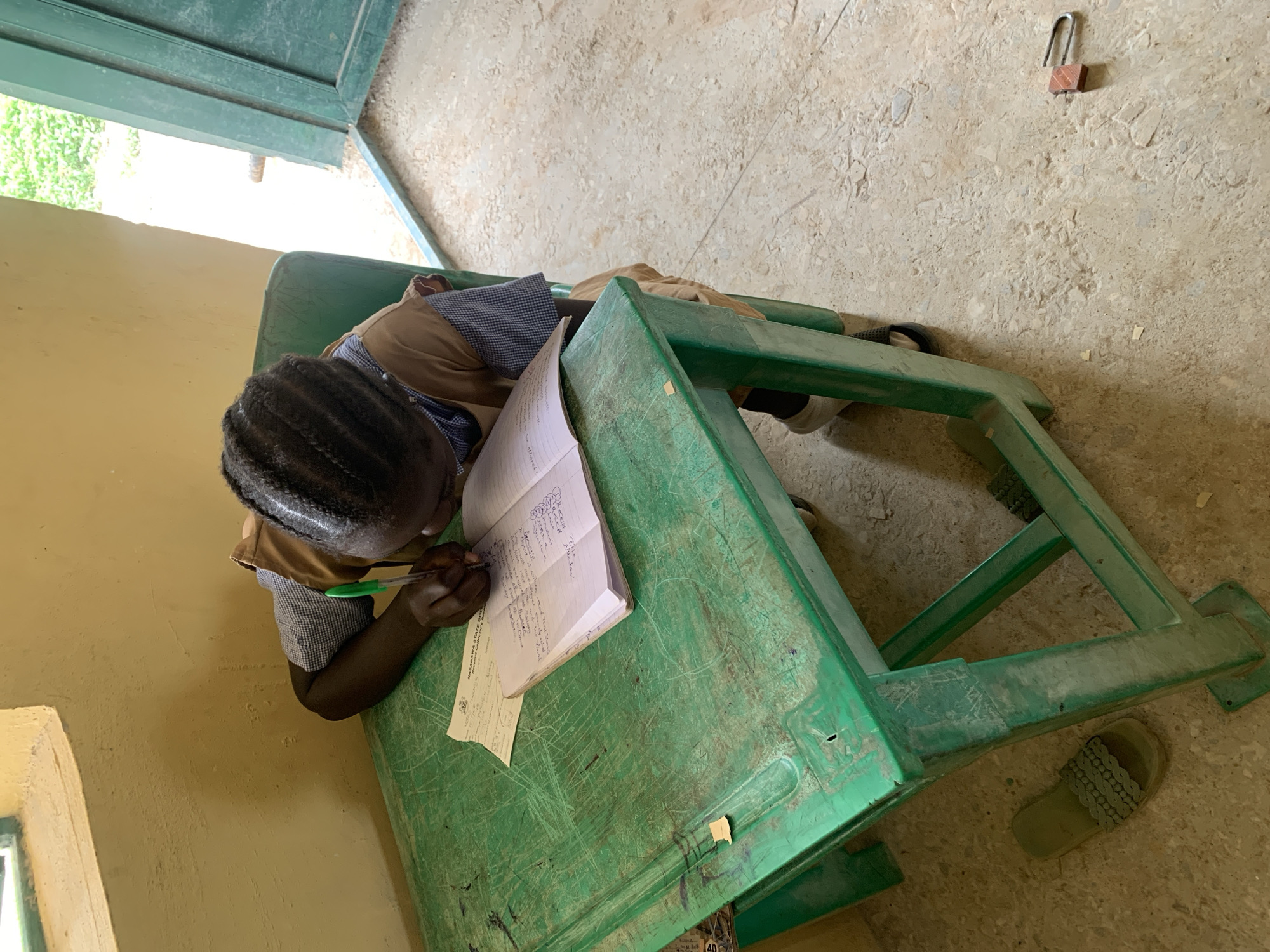 The media file shows a young person, likely a student, writing in a notebook at a green plastic desk. The student is wearing a brown shirt over a blue and white checkered shirt. Their hair is braided. The desk is worn and scratched, with some papers on it, including what appears to be a form with text. A green door is partially visible in the background, opening to an area with greenery outside, suggesting it might be daytime. The floor is concrete and appears to be in a somewhat unfinished or rustic setting, possibly a classroom in Akwanga, Nigeria, given the location context. A single sandal is on the floor next to the desk. A small, open padlock is visible on the wall in the background. The scene appears to be one of focused study.