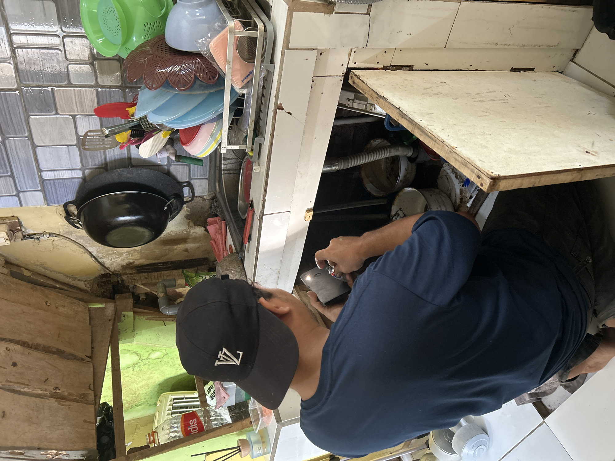 In a cluttered kitchen, a person wearing a dark blue t-shirt and a black baseball cap with a white logo is hunched over, working on something held in their hands. The environment appears to be an indoor space, possibly a home kitchen, with a sink area visible. Dishes and kitchen utensils are stacked on a rack next to the sink. The cabinetry beneath the sink is open, revealing pipes and containers. The overall scene suggests a repair or maintenance task is being performed in a somewhat disorganized setting. The image is taken from behind and to the side of the individual.