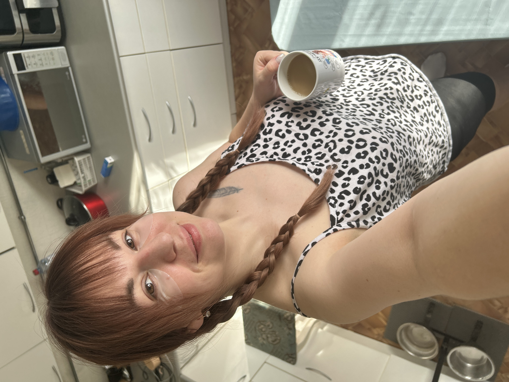 This image depicts a female in a domestic kitchen environment, located in the city of Фоки, Russia. The subject, an adult female with reddish-brown hair styled with bangs and two braids, is seen from the chest up, holding a white mug with a light brown liquid in her right hand. She is wearing a white and black leopard-print tank top and dark leggings, with clear hydrogel patches applied beneath both eyes. A small feather tattoo is visible on her left shoulder. She is smiling and looking directly at the camera, indicating a selfie.
The background shows a kitchen with white upper and lower cabinets and a light grey countertop. Visible appliances include a silver LG microwave oven and another silver appliance, possibly an air fryer, positioned on the countertop. A red pot with a lid, a clear plastic bottle, a white basket containing items, and a small blue box are also on the counter. The flooring is light brown, possibly laminate or tile. On the floor to the lower left, two empty stainless steel pet bowls rest on a dark mat. A light blue textile object is visible on the floor to the lower right. Natural light casts shadows across the scene.