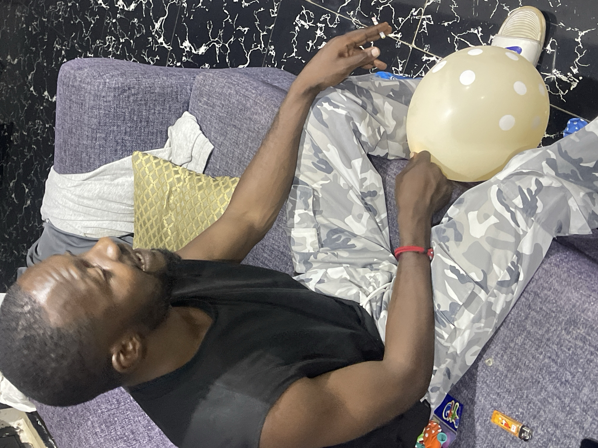 A dark-skinned man with a beard and short hair is sitting on a purple couch, holding a beige balloon with white polka dots. He is wearing a black sleeveless shirt and camouflage pants. His right arm is extended, and he is holding a cigarette between his index and middle fingers. A lighter is on the couch next to him. The floor is black and white marble. The image appears to have been taken indoors. The man's gaze is directed away from the camera. The overall mood is casual.