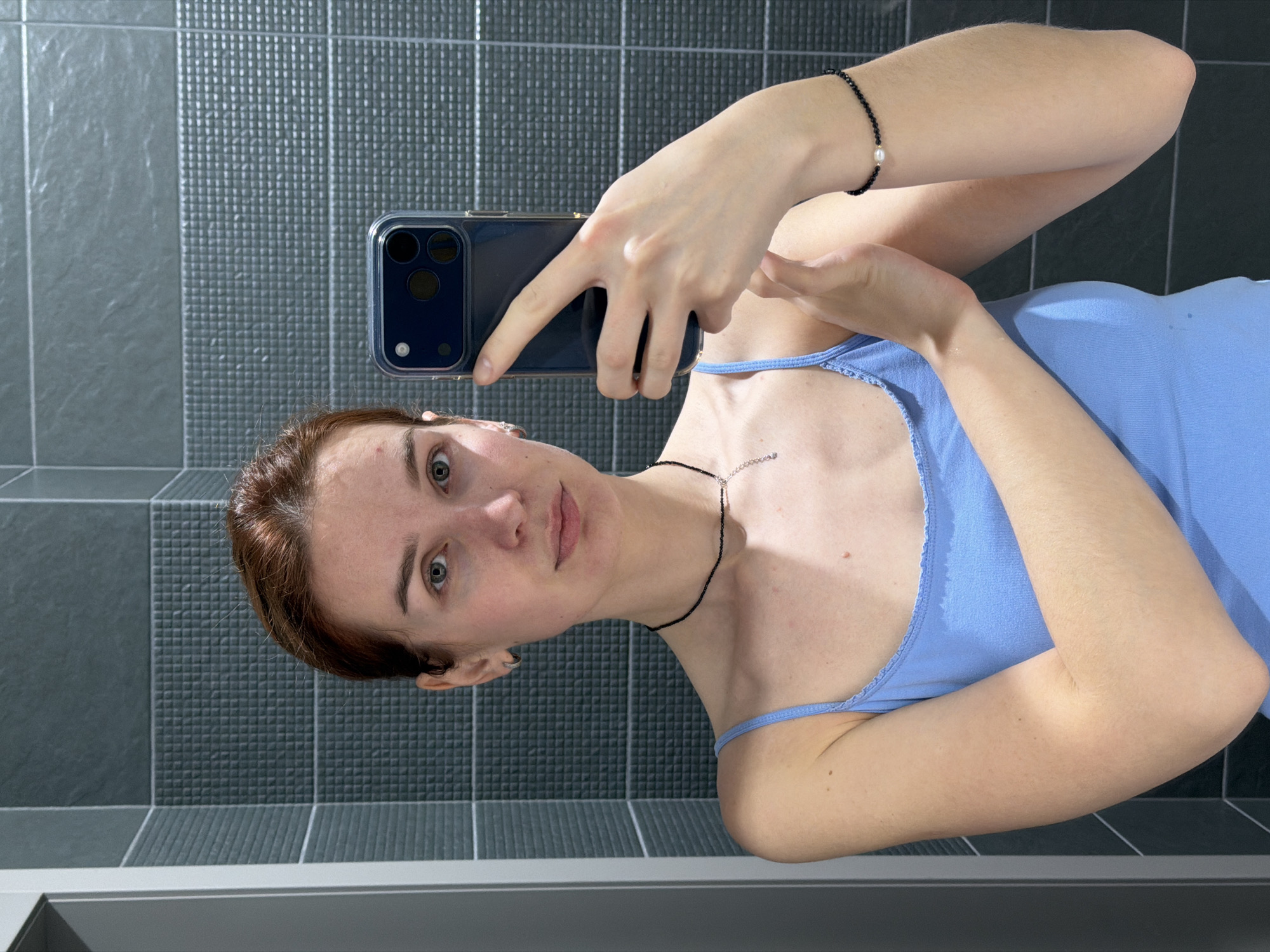 A selfie captured in a bathroom with gray tiled walls. A person with auburn hair and blue eyes is holding a blue smartphone to take the picture. They are wearing a light blue tank top and two necklaces: one black beaded, and one silver with small stones. A black beaded bracelet with a pearl is on the wrist of the arm holding the phone. The location is potentially a bathroom located in the city of Chaikovsky, Russia.