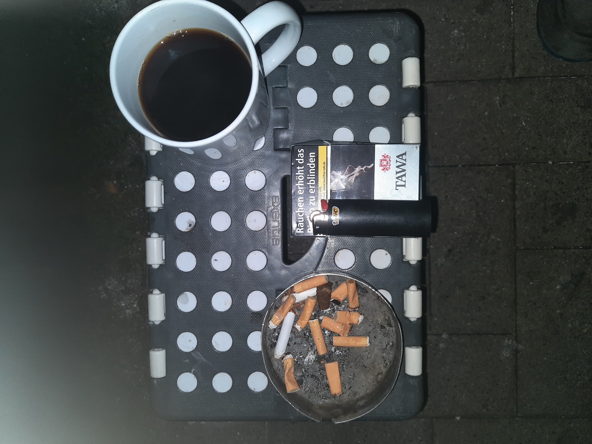 In the city of Celle, Germany, an overhead shot depicts a small, dark grey portable stool with a textured surface featuring a pattern of circular indentations. Resting on the stool are several objects. To the left, a round, metallic ashtray is filled with numerous cigarette butts, some with orange filters and others with white filters, along with ash and debris. Next to the ashtray, a black lighter lies horizontally. To the right of the lighter, a pack of "TAWA" cigarettes is positioned vertically, displaying a warning label in German that reads "Rauchen erhöht das zu erblinden" (Smoking increases the risk of blindness) and an image of smoke. To the right of the cigarette pack, a white ceramic mug is filled with dark liquid, presumably coffee. The stool is placed on a dark, textured surface, possibly pavement or an outdoor floor. The lighting suggests it is nighttime or a dimly lit environment.