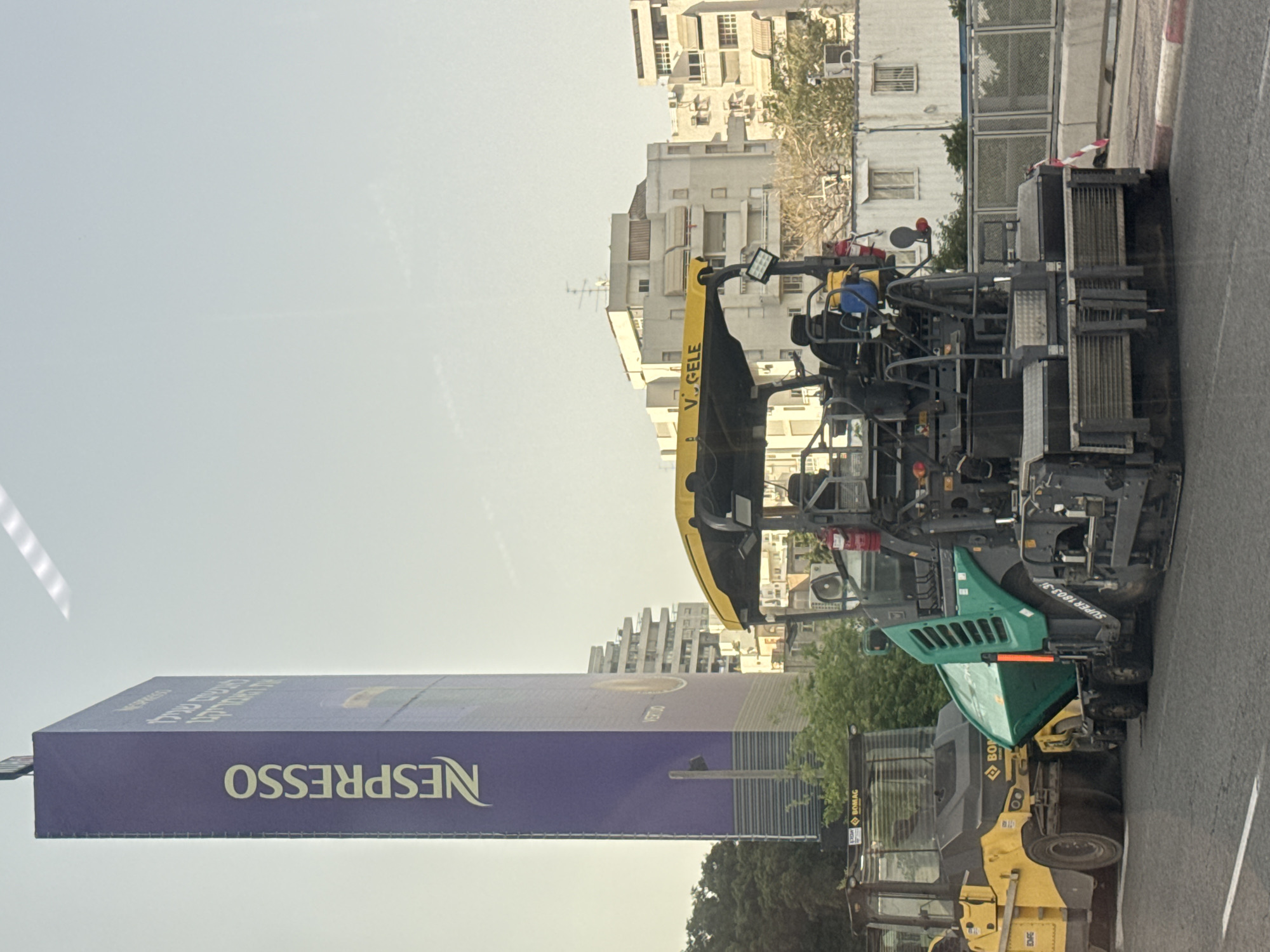 The image shows an urban scene in Tel Aviv, Israel. A large "Nespresso" billboard stands prominently on the left. In the background, residential buildings are visible under a hazy sky. A road construction vehicle, specifically an asphalt paver, and a BOMAG road roller are positioned on the right side of the frame. The paver has a Vögele logo, and the Bomag roller is yellow and black. The vehicles appear to be stationary, likely involved in road maintenance or construction activities. The view suggests the photo was taken from within another vehicle looking out onto the scene.