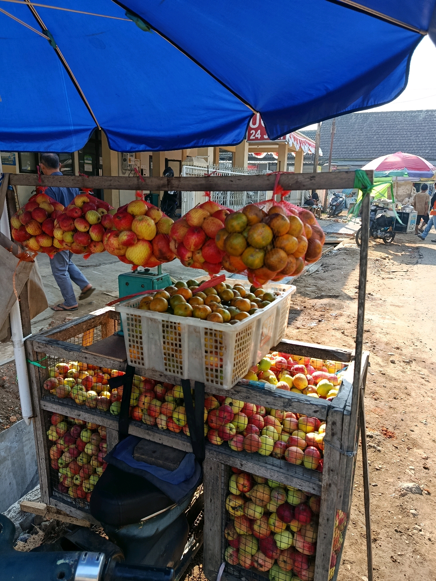A roadside fruit stand is set up under a large blue tarp in Bululawang, Indonesia. The stand displays a variety of fresh fruits, predominantly apples and oranges. The apples are bagged in yellow mesh sacks, with several bags hanging from the wooden structure of the stand. Below the hanging bags, a white plastic crate is filled with oranges. Two wooden crates, each with a wire mesh front, are filled with apples. The lower crate also contains some oranges. A black motorcycle seat is visible in the foreground. In the background, a person walks past the stand, and other vehicles and market stalls can be seen, suggesting a busy street scene. The ground is dirt and gravel, and the lighting indicates it is daytime.