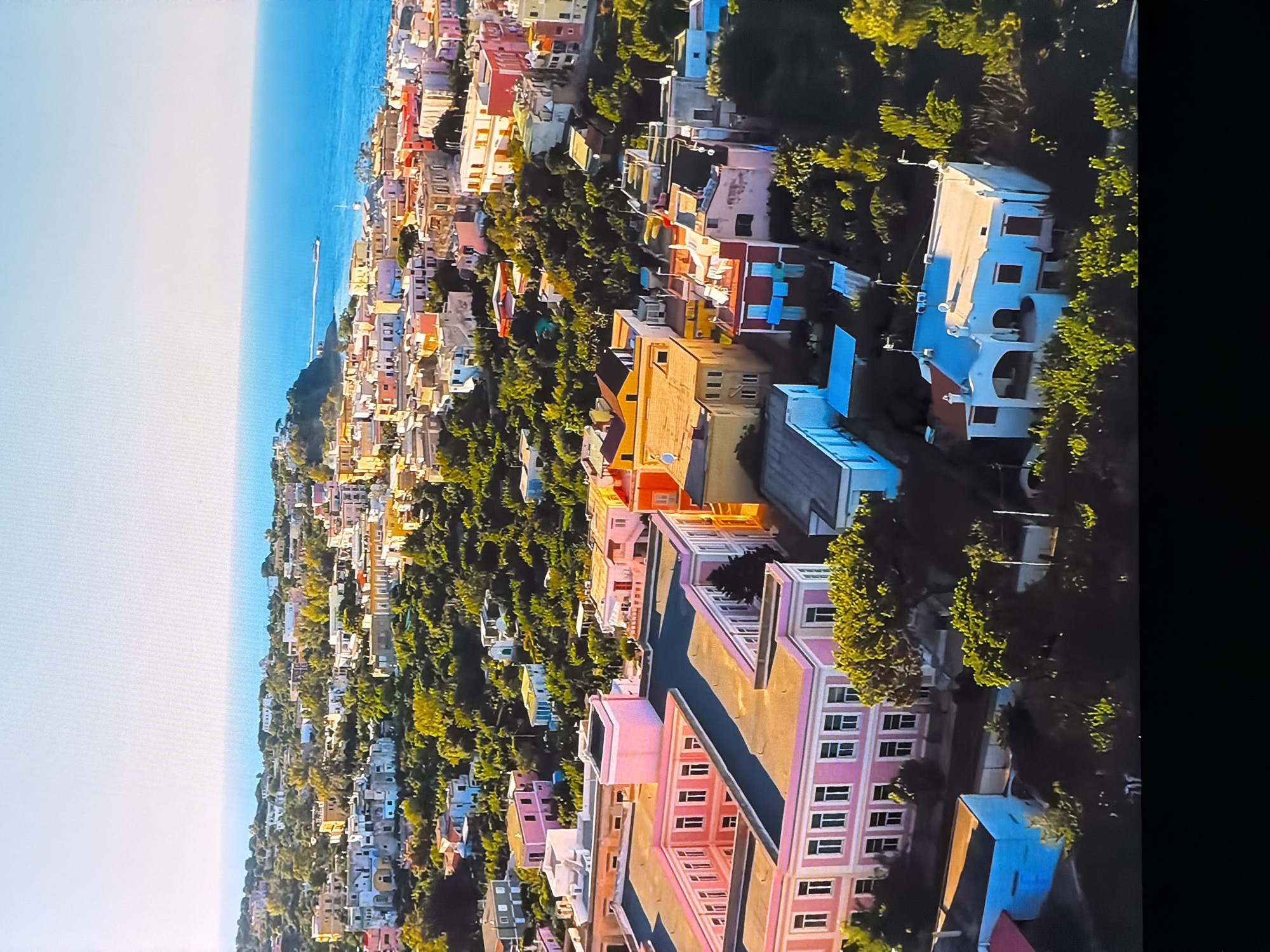 The image is an aerial view of a coastal town with colorful buildings nestled among lush green trees. The buildings are of varying sizes and architectural styles, with many featuring pastel-colored facades, terracotta roofs, and balconies. The town is situated on a hillside that slopes down towards a bright blue sea, with a clear, light blue sky above. In the foreground, a large, pink, U-shaped building with a dark roof stands out, surrounded by trees. Further into the town, smaller residential buildings and some larger structures are visible. The overall impression is one of a vibrant, sunny, Mediterranean-style coastal community. There are no people visible in the image. The scene does not depict Chirchik, Uzbekistan, as it is a coastal town and Uzbekistan is a landlocked country.