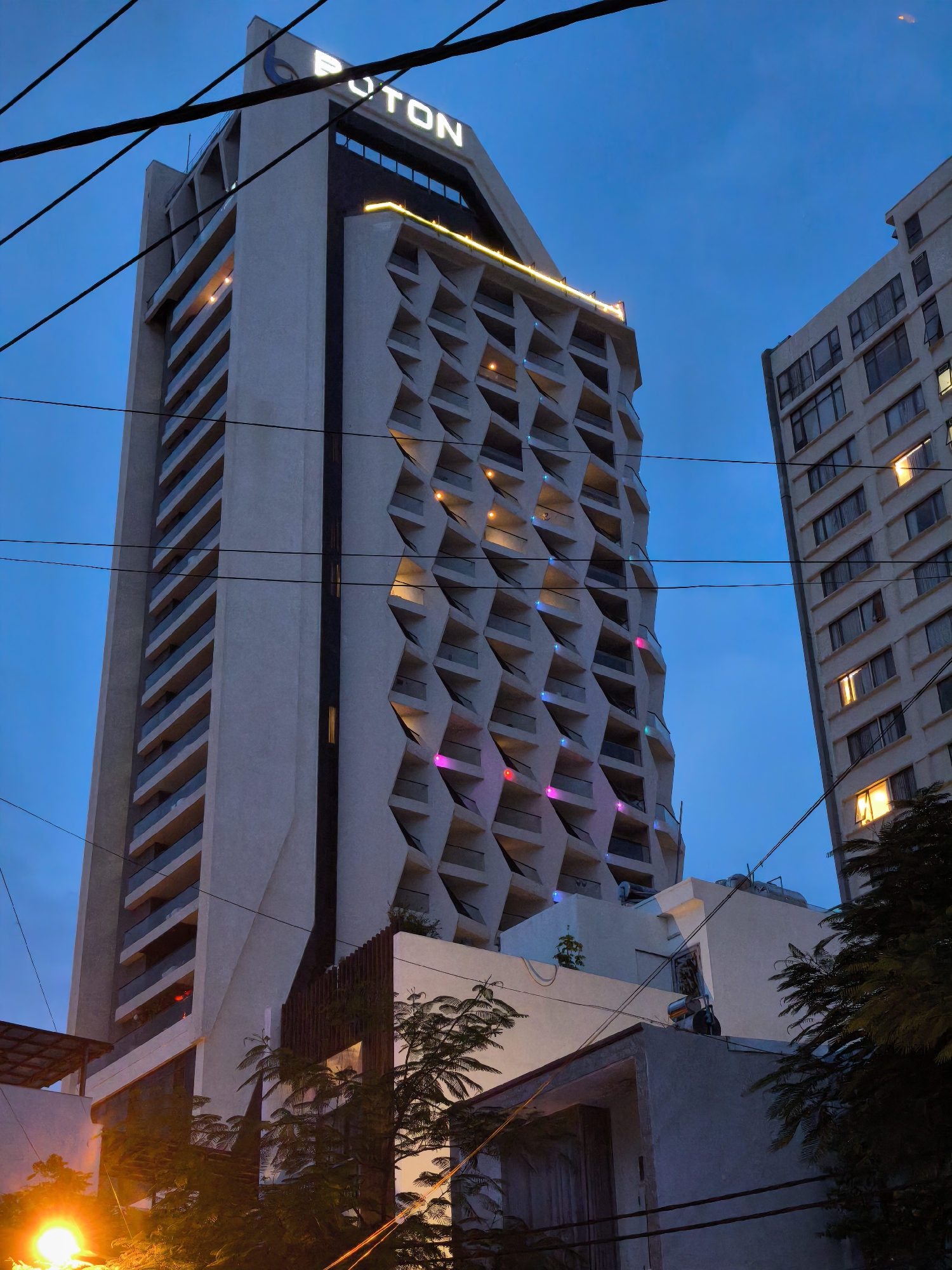 The image depicts a twilight scene in Bac Nha Trang Ward, Vietnam, focusing on modern high-rise buildings. The central subject is a tall, irregularly shaped building with a facade featuring a repeating diamond pattern, accented by colorful lights on multiple levels. Above this building, the "PUTON" logo is visible in illuminated white letters against a dark blue sky. To the right, another, more conventionally designed, tall building with numerous windows, some illuminated, stands partially obscured. The foreground and mid-ground are dominated by the lower portions of these structures, along with some trees and what appears to be a lower building or part of a complex, partially silhouetted against the evening sky. Overhead, numerous power lines crisscross the frame, adding a layer of urban complexity. The lighting suggests it is either early evening or late dusk, with artificial lights from the buildings and a bright street lamp in the lower left corner illuminating parts of the scene. No people are visible.