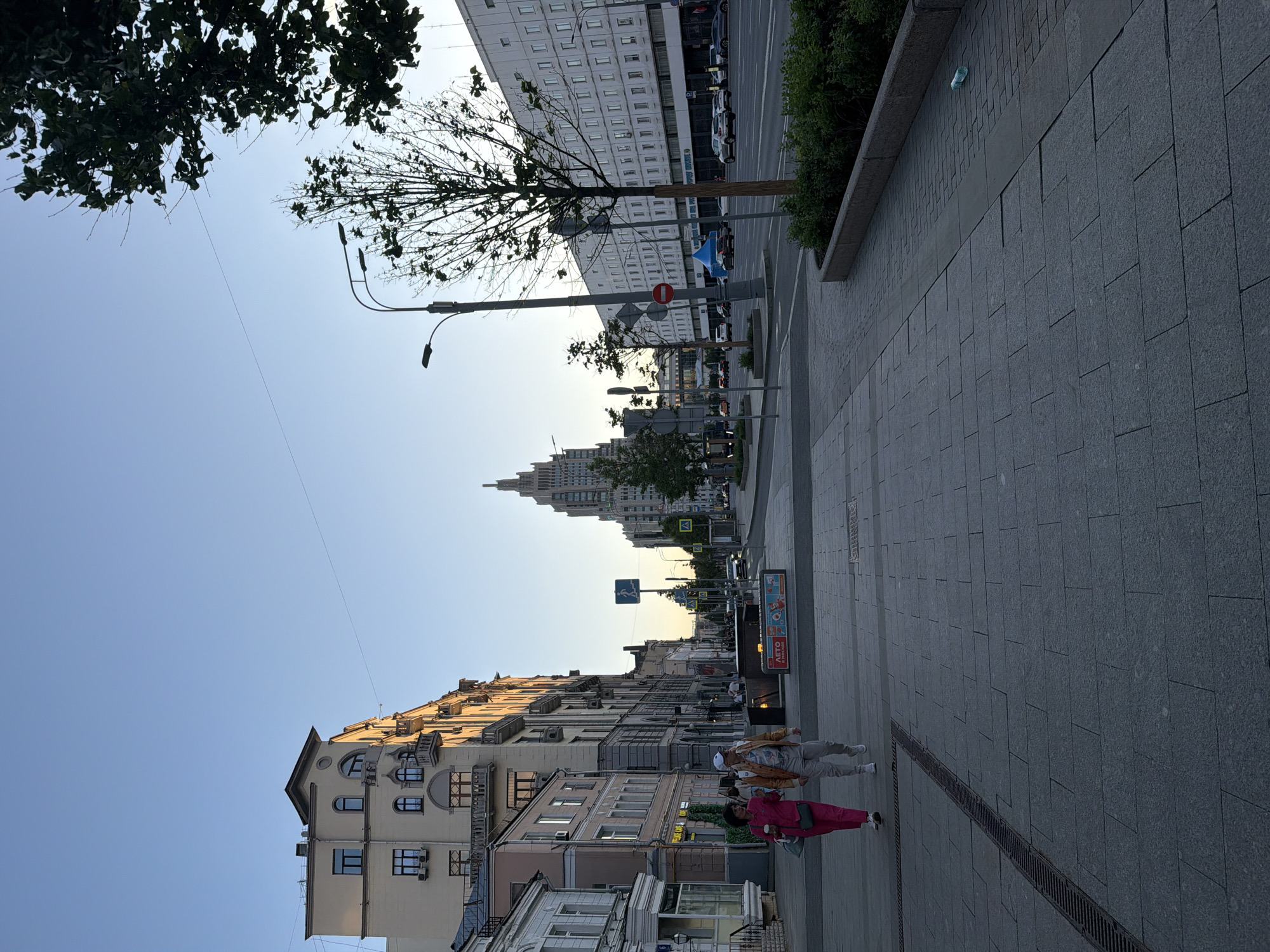 The image shows a street scene in Moscow, Russia. The perspective is along a wide sidewalk paved with grey stones, bordered by buildings on either side. The architecture includes a mix of old and modern styles, with some buildings showing ornate details and others being more utilitarian in design. In the distance, a prominent skyscraper with a spire is visible, likely one of the Seven Sisters, Stalinist skyscrapers that are landmarks of Moscow.
Several pedestrians are walking on the sidewalk. The street is visible between the buildings, with cars parked along the sides. There are street signs and traffic lights, indicating an urban environment. Trees are sparsely planted along the street, and there is some greenery in planters. The sky is clear and light, suggesting either morning or late afternoon.