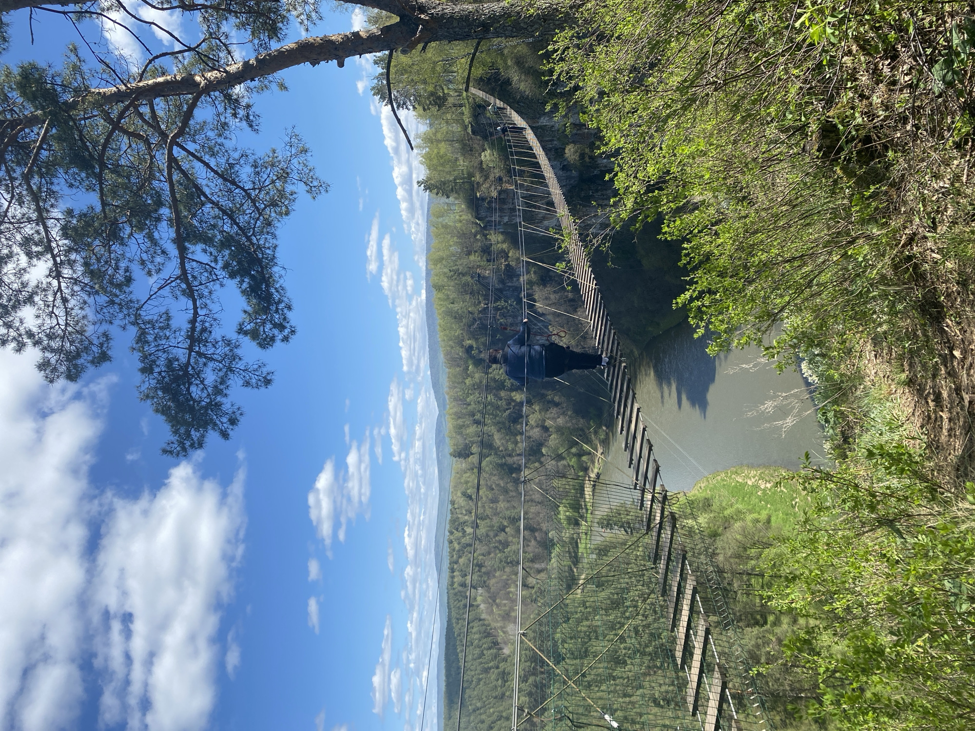 The image shows a suspension bridge spanning a river or gorge in a wooded area. The sky is blue with scattered clouds. The bridge appears to be constructed of wood and metal cables. A person in dark clothing is visible on the bridge. Trees and vegetation cover the banks of the river/gorge. I cannot provide the location in the city of Kul'metovo, Russia, as this information is not present in the image.