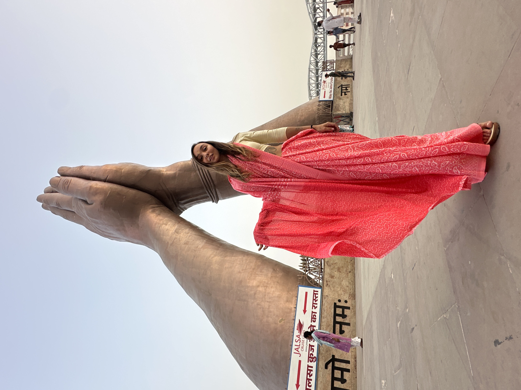 A vertical full shot depicts a woman lying on a large sculpture of praying hands in Mughalsarai, India. The woman is fair-skinned, with long brown hair, wearing a gold top and a bright pink saree with a white pattern. The giant hands are bronze in color. In the background, other pedestrians are walking on the same platform. A sign indicates "JALSA Cruises" in both English and Hindi. The sky is a light, hazy blue.