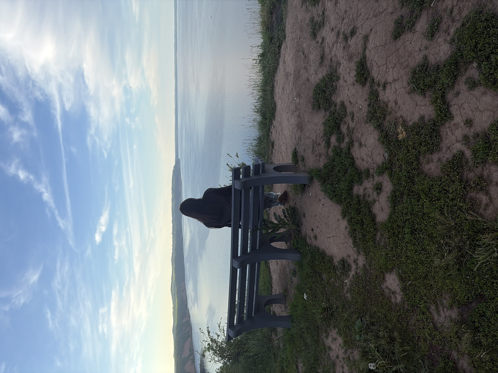 The image captures a serene outdoor scene, rotated so the horizon is vertical. When correctly oriented, a person with long, dark hair is seated on a dark grey park bench, viewed from behind, looking out over a vast body of water. The bench rests on an elevated natural landscape, likely a cliff edge or high bank, characterized by dry, cracked earth interspersed with patches of green grass and small bushes.
The individual is looking towards the expansive body of water, which stretches to a distant landmass or horizon line. The sky above is a vibrant blue, filled with delicate, wispy white clouds, indicating clear weather and soft daylight, possibly late afternoon or early morning. The overall atmosphere conveys peace and contemplation, with the solitary figure enjoying the expansive view. There is no visible text in the image. The setting suggests a natural coastal or large river/lake environment in the provided context of Костоваты, Russia.