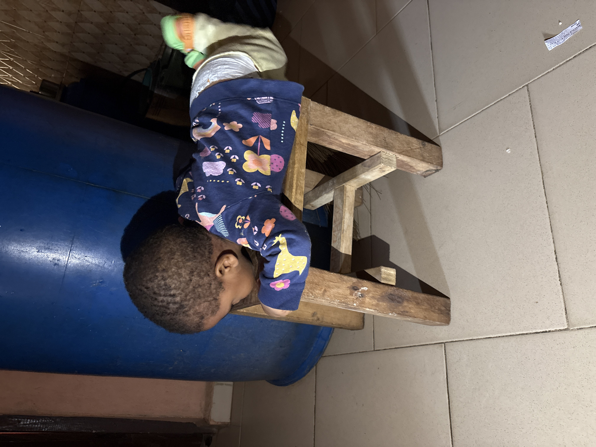 The image shows a young child lying face down on a small wooden stool. The child is wearing a dark blue long-sleeved shirt with a colorful pattern of flowers and shapes, and a diaper. The child's head is turned to the side, resting on the stool. One of the child's feet, wearing a green sock, is visible extending off the stool. The stool is positioned on a tiled floor. In the background, there is a large blue cylindrical object, possibly a barrel or tank, and a tiled wall. The lighting suggests an indoor setting at night or in a dimly lit area. The scene appears to be in a residential or utilitarian space, possibly within the city of Onitsha, Nigeria, given the tiled flooring and background elements.